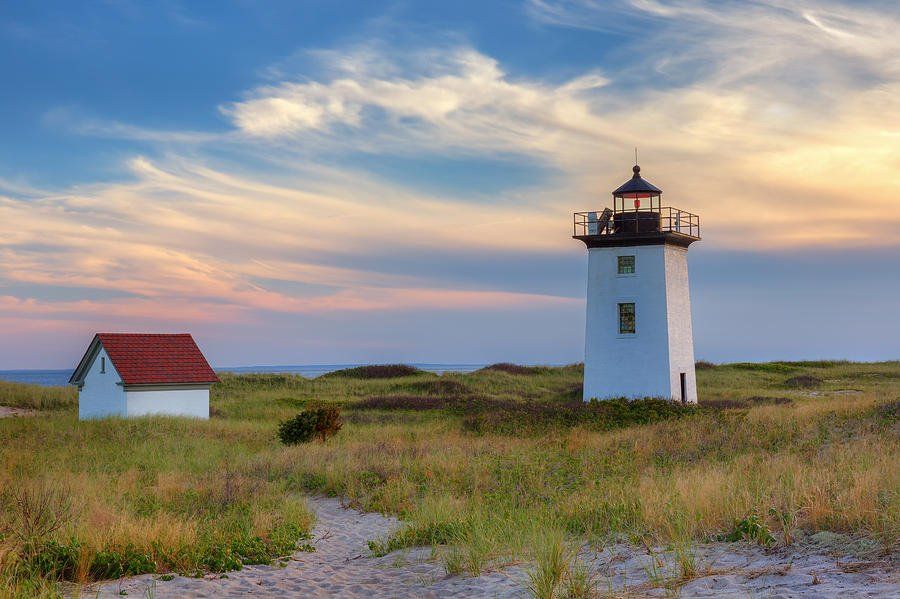 White lighthouse and small red-roofed building on a grassy dune under a colorful sky at sunset.