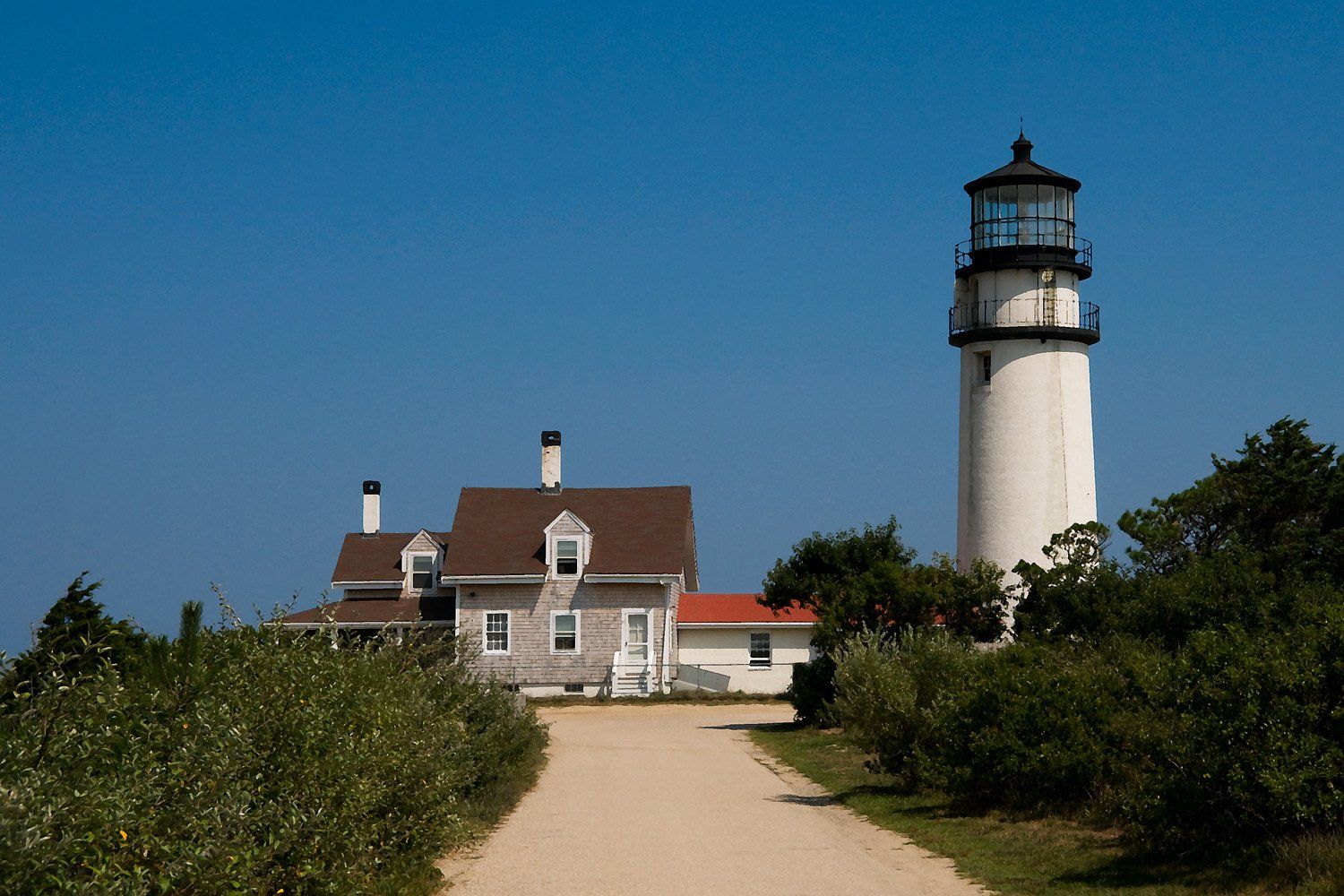 White lighthouse and adjacent house against a clear blue sky, a path leading towards them.