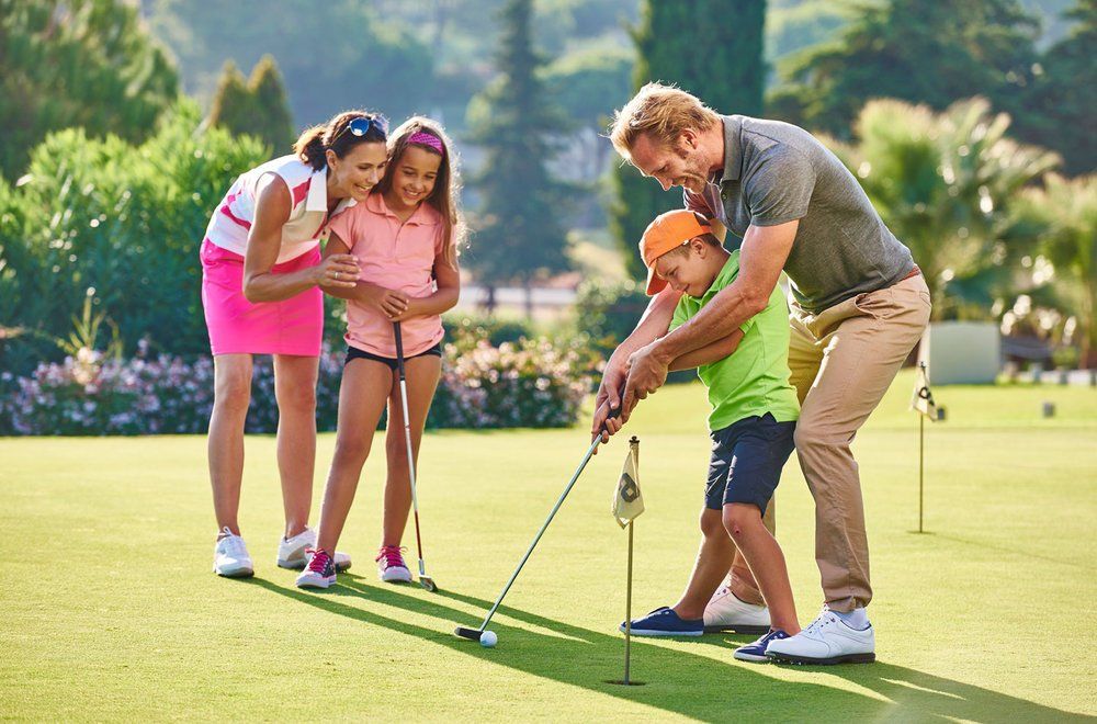 Family golfing: father coaching son, mother and daughter watching on green grass. Sunny day.