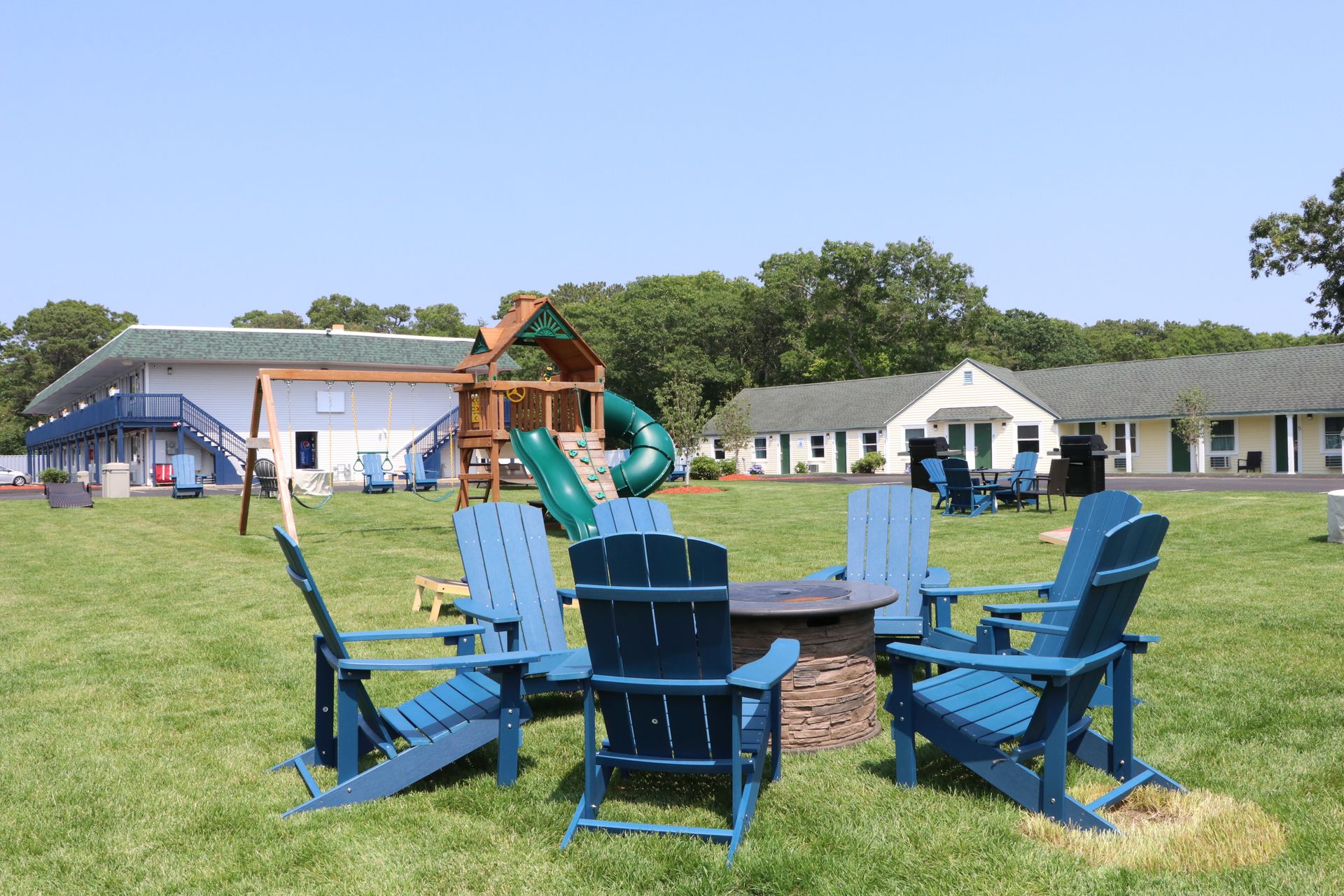 Blue Adirondack chairs surround a fire pit in a grassy yard, motel buildings in the background.