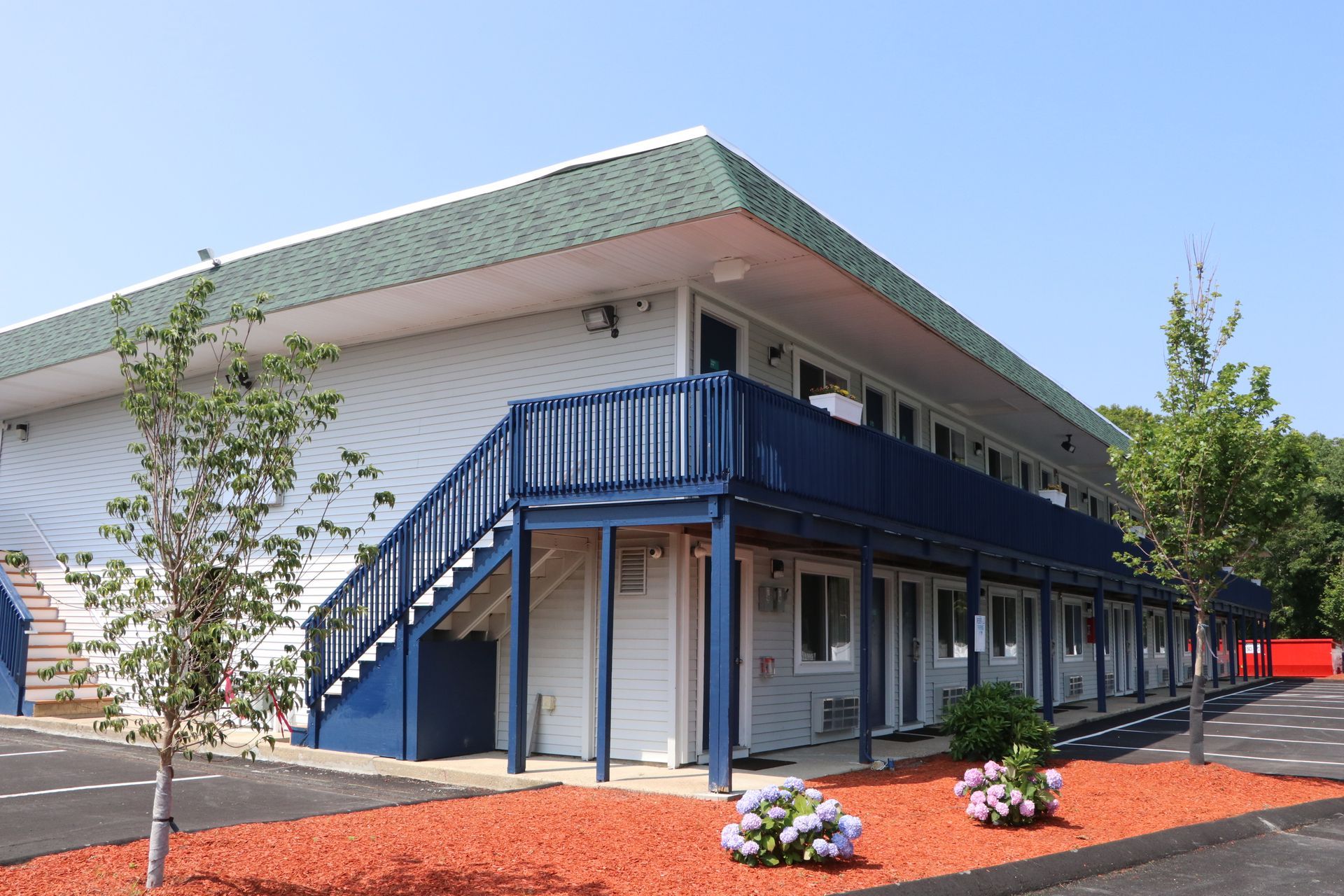 Two-story motel with white siding, blue trim, and green roof, red mulch, and blue stairs. Sunny day.