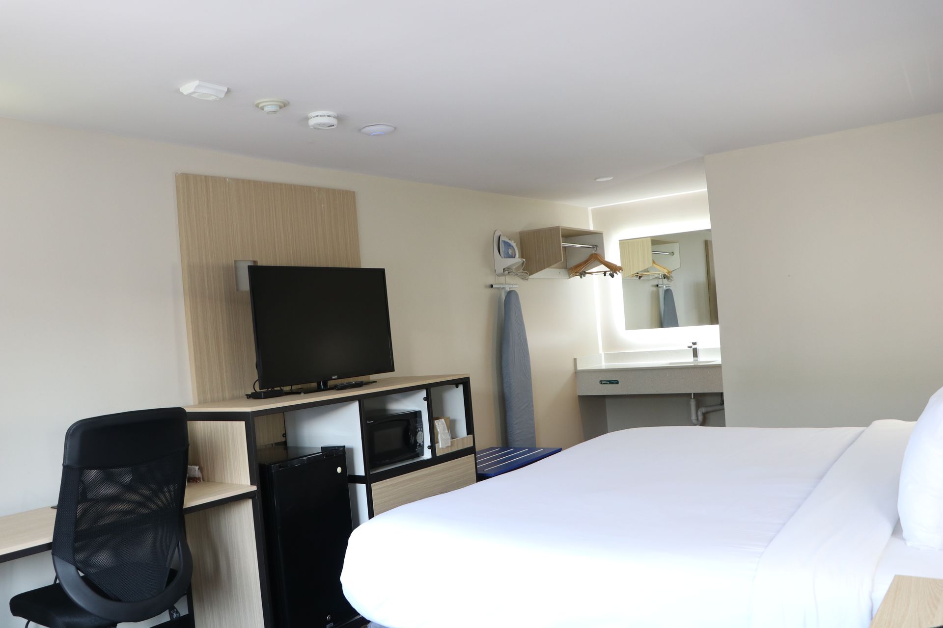 Hotel room with a bed, TV, desk, and bathroom visible in the background. Bright, white walls.