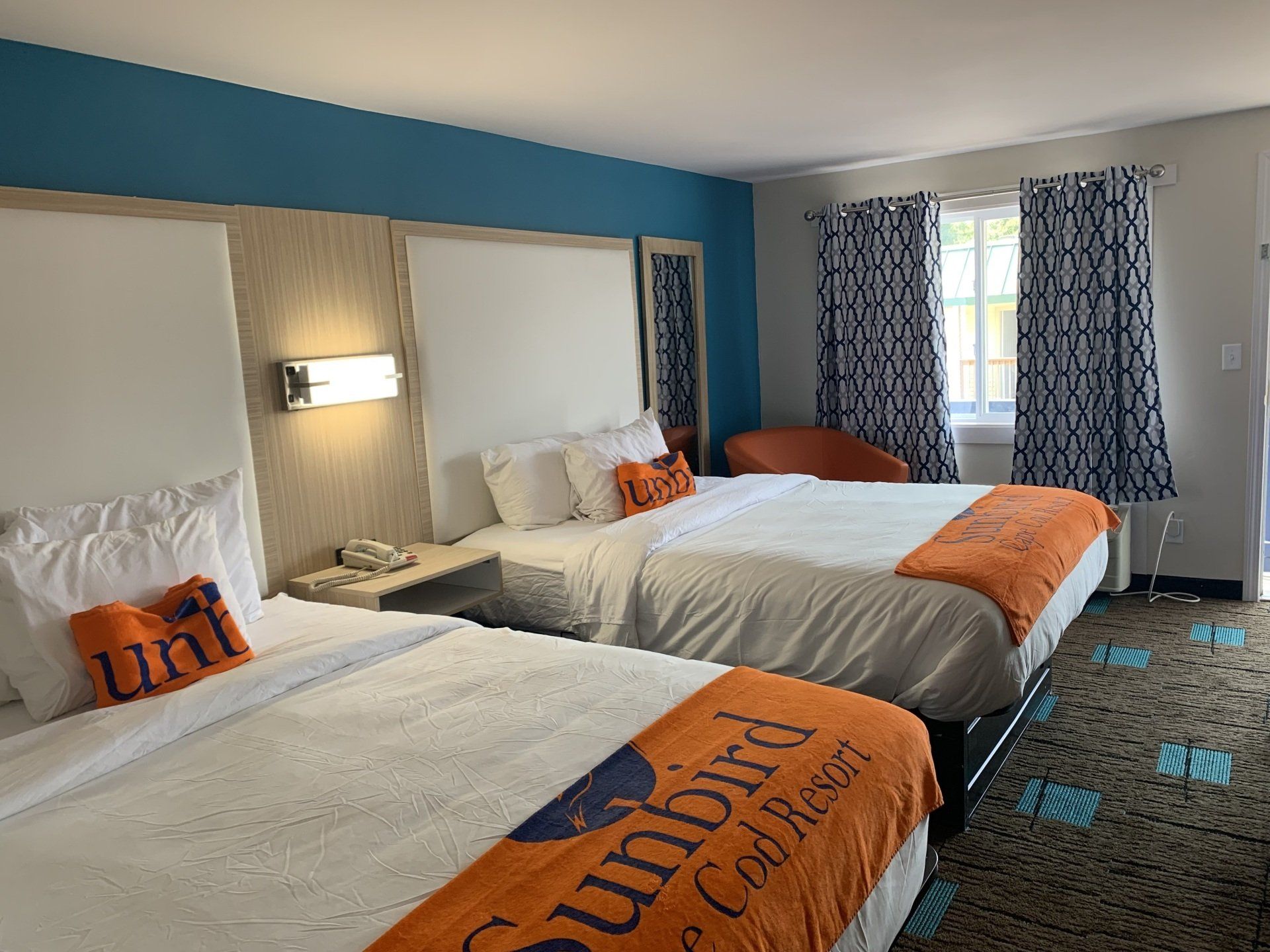 Hotel room with two queen beds, orange and blue accents, patterned carpet and curtains.