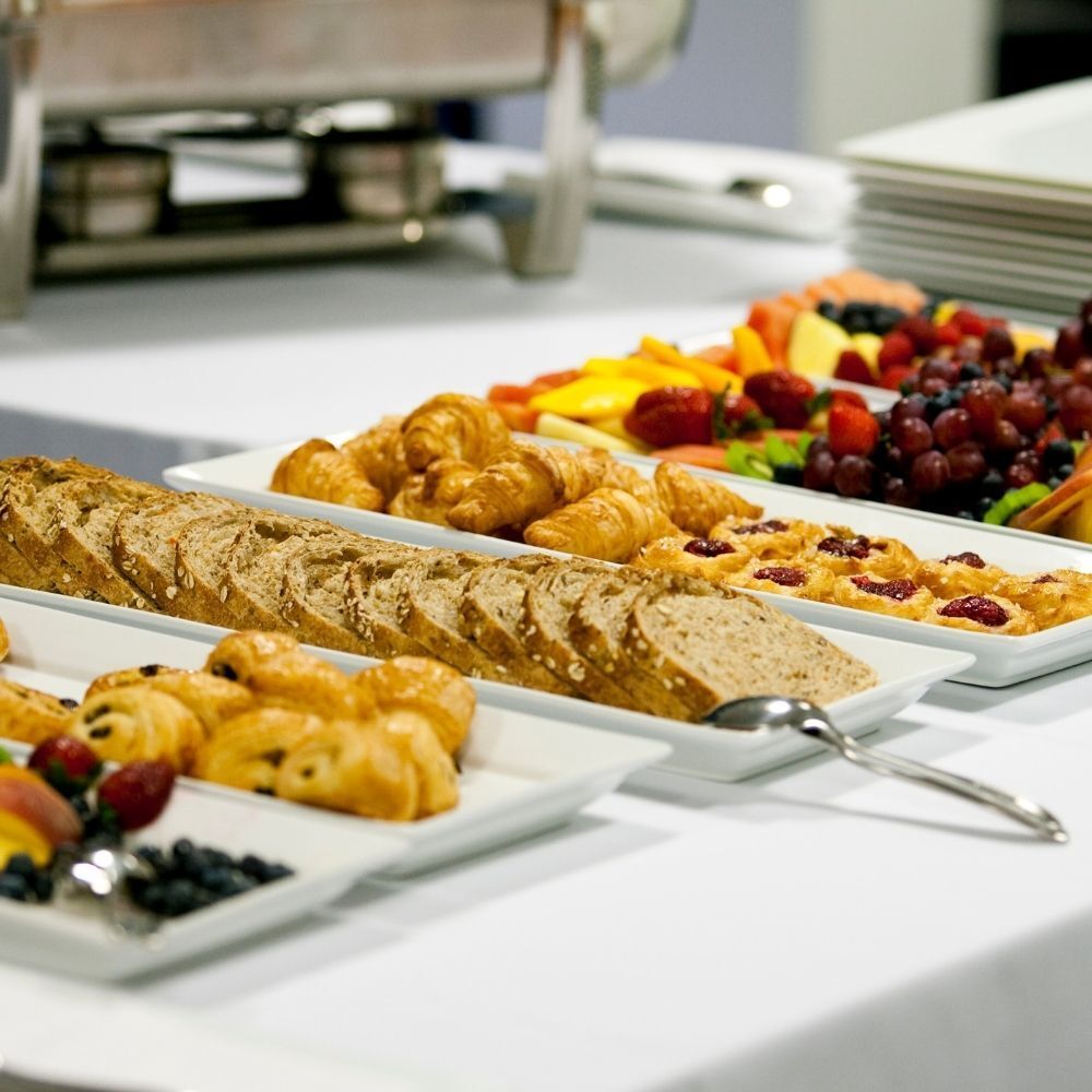 Buffet table with bread, croissants, fruit, and chafing dishes on a white tablecloth.