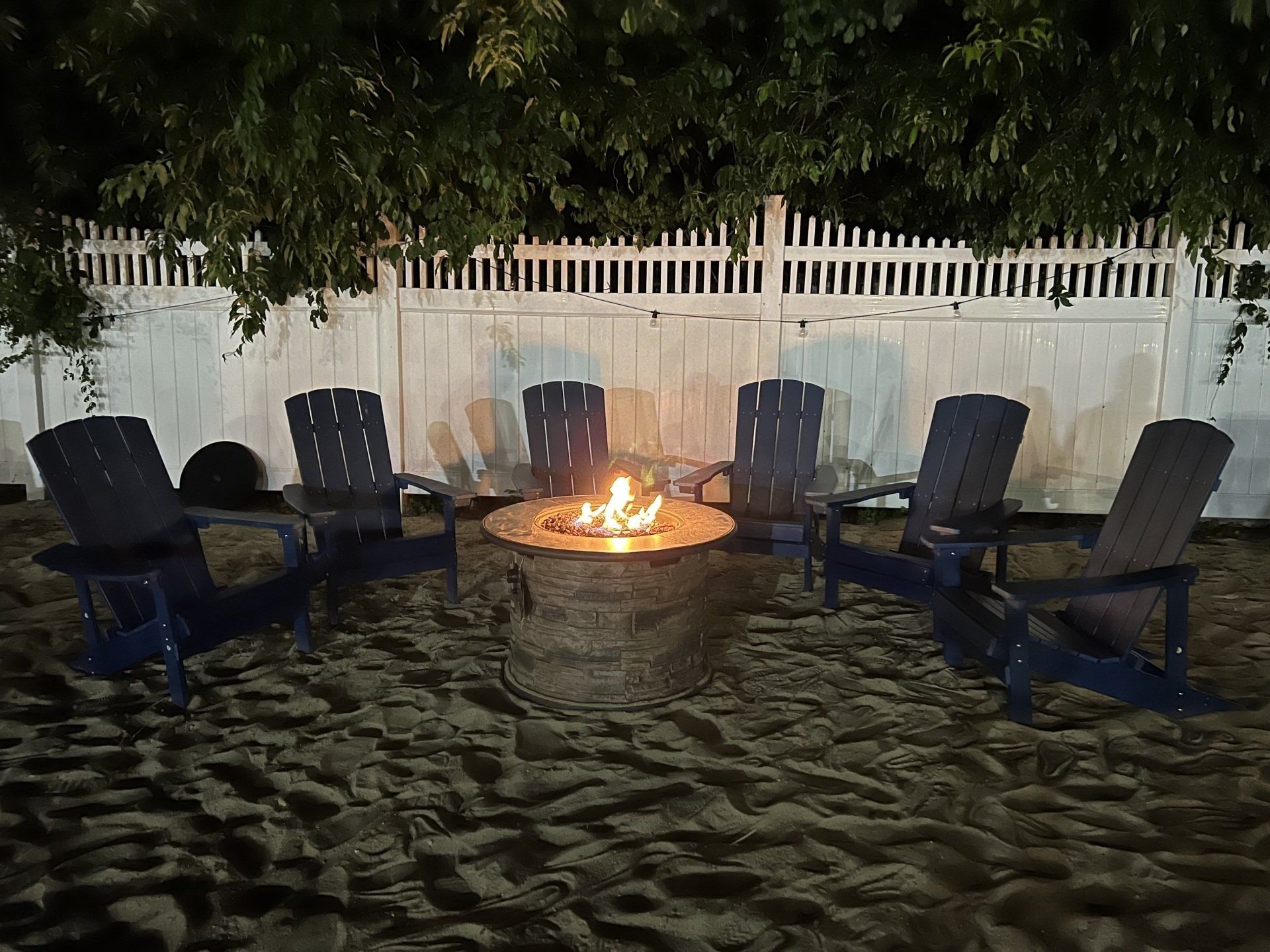 Six dark blue chairs around a fire pit on sand, beneath a white fence at night.