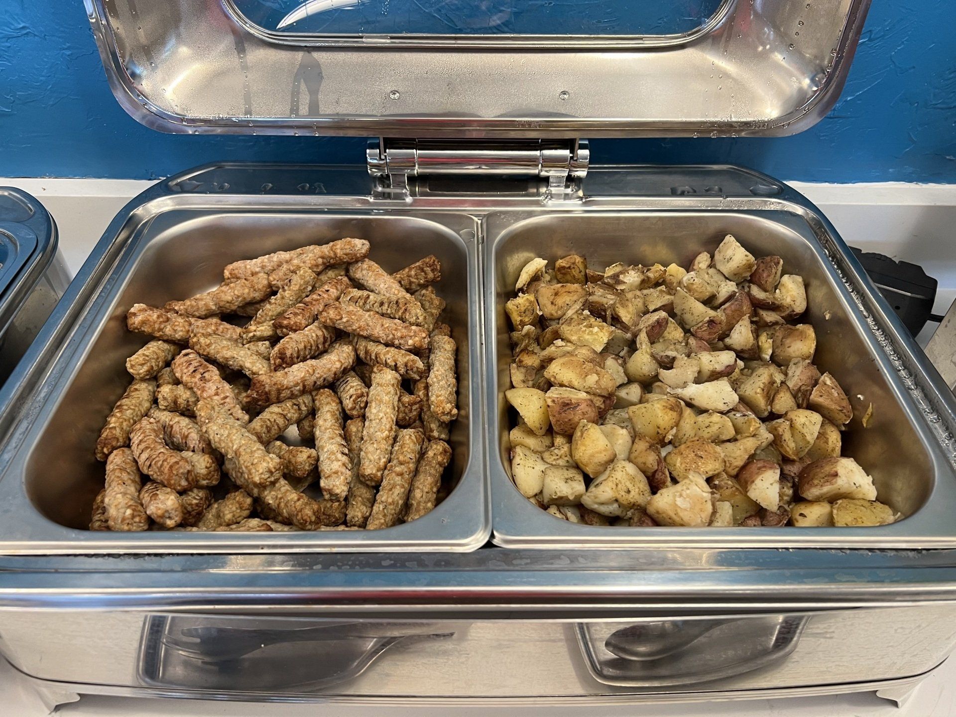 Breakfast buffet with sausage links and diced potatoes in stainless steel warming trays.