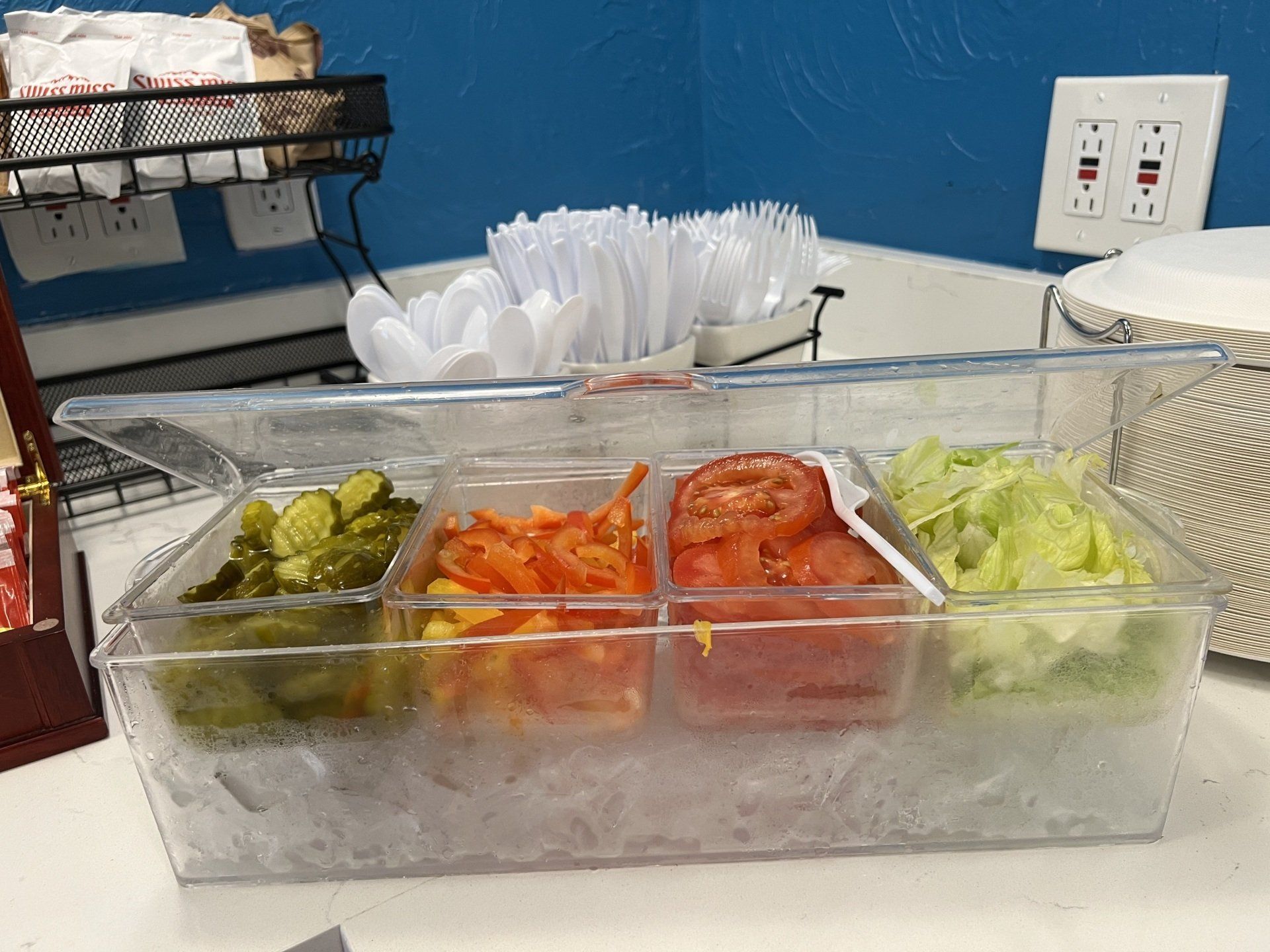 A clear container holding pickles, carrots, tomatoes, and lettuce; ready for sandwiches.