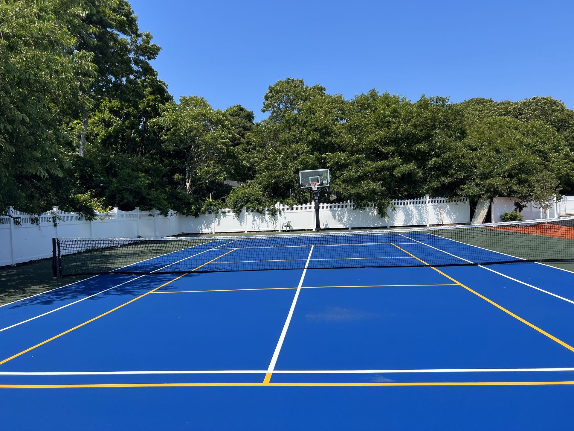 Blue tennis court with white lines and net, basketball hoop in the background, surrounded by white fence and trees.