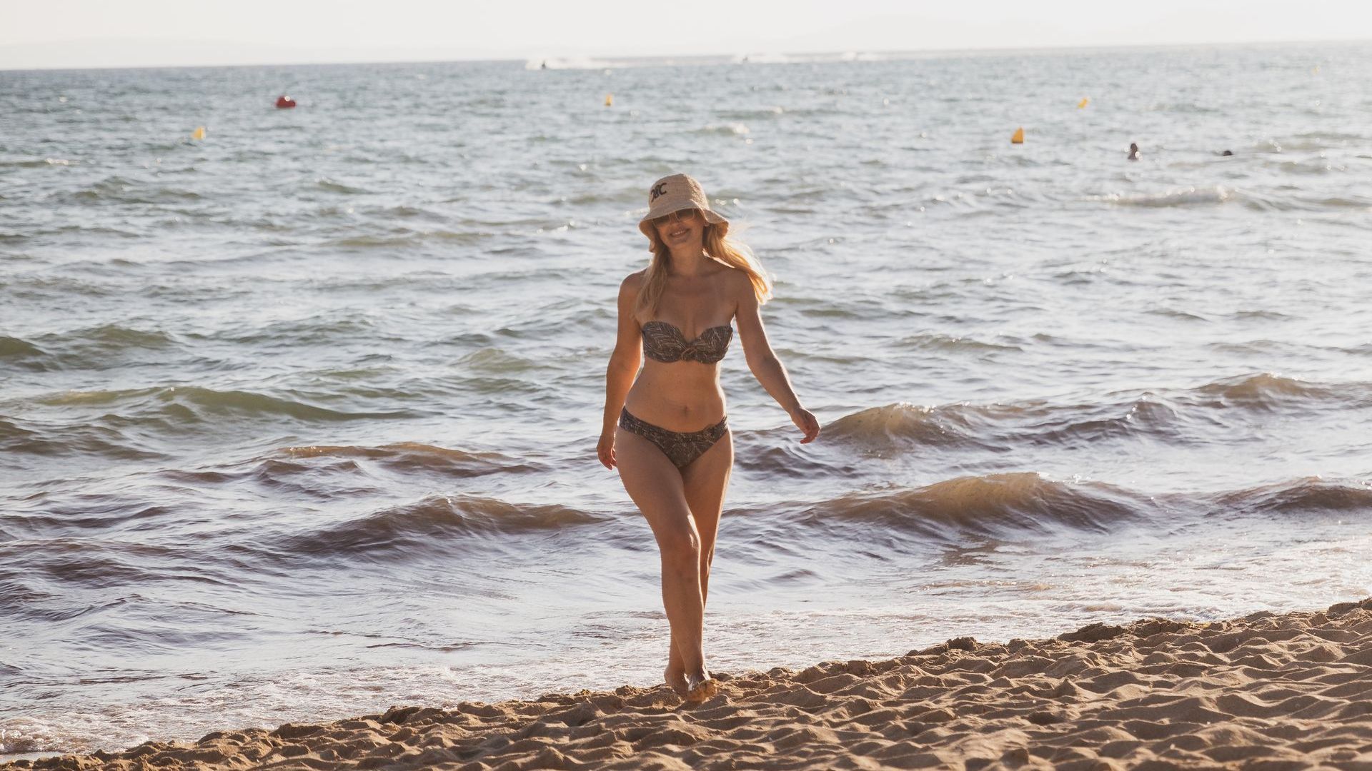 Eine Frau in Bikini und Strohhut geht am Strand spazieren.