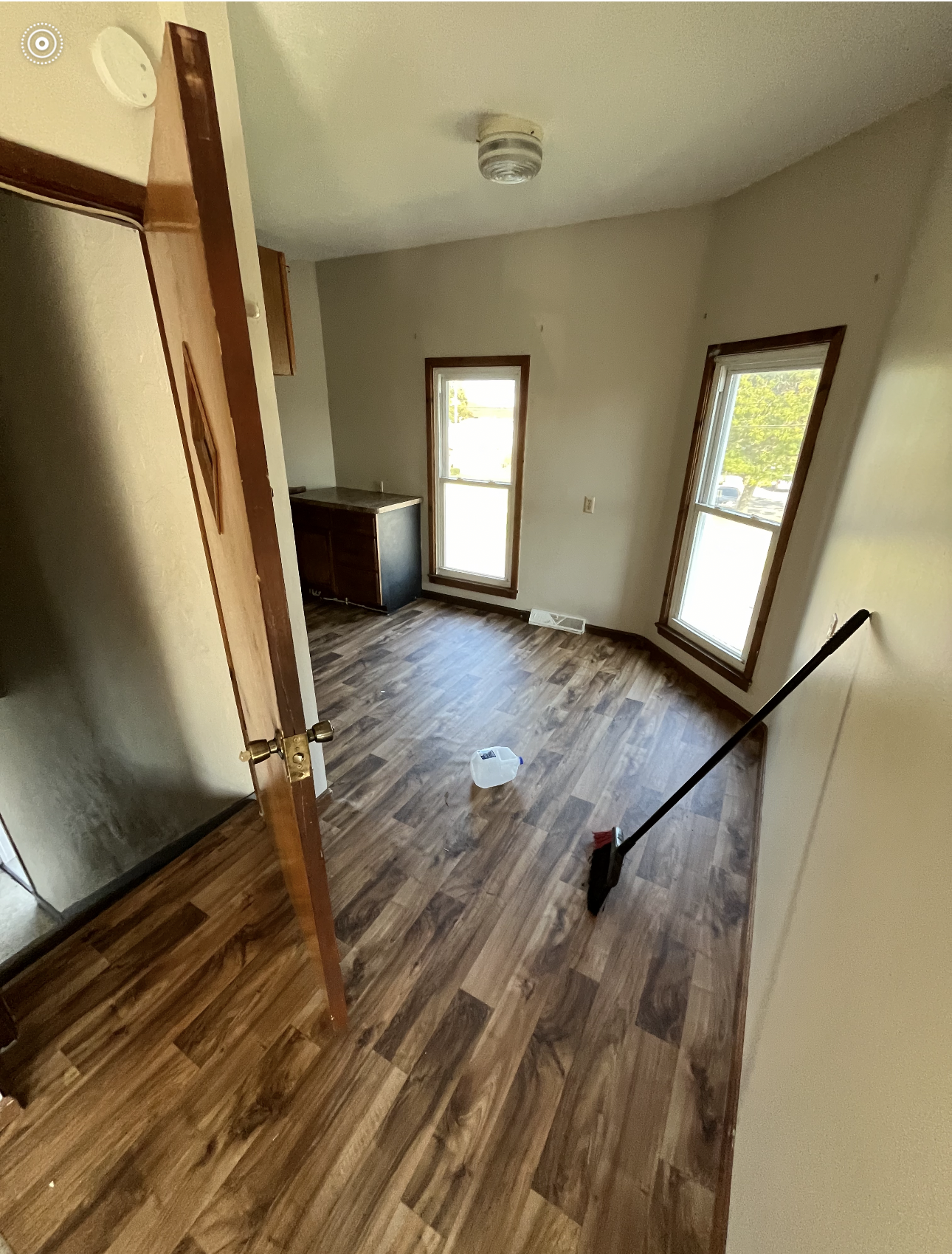 Room with wood-look flooring, two windows, and dark storage unit. A broom and a robotic vacuum are visible.