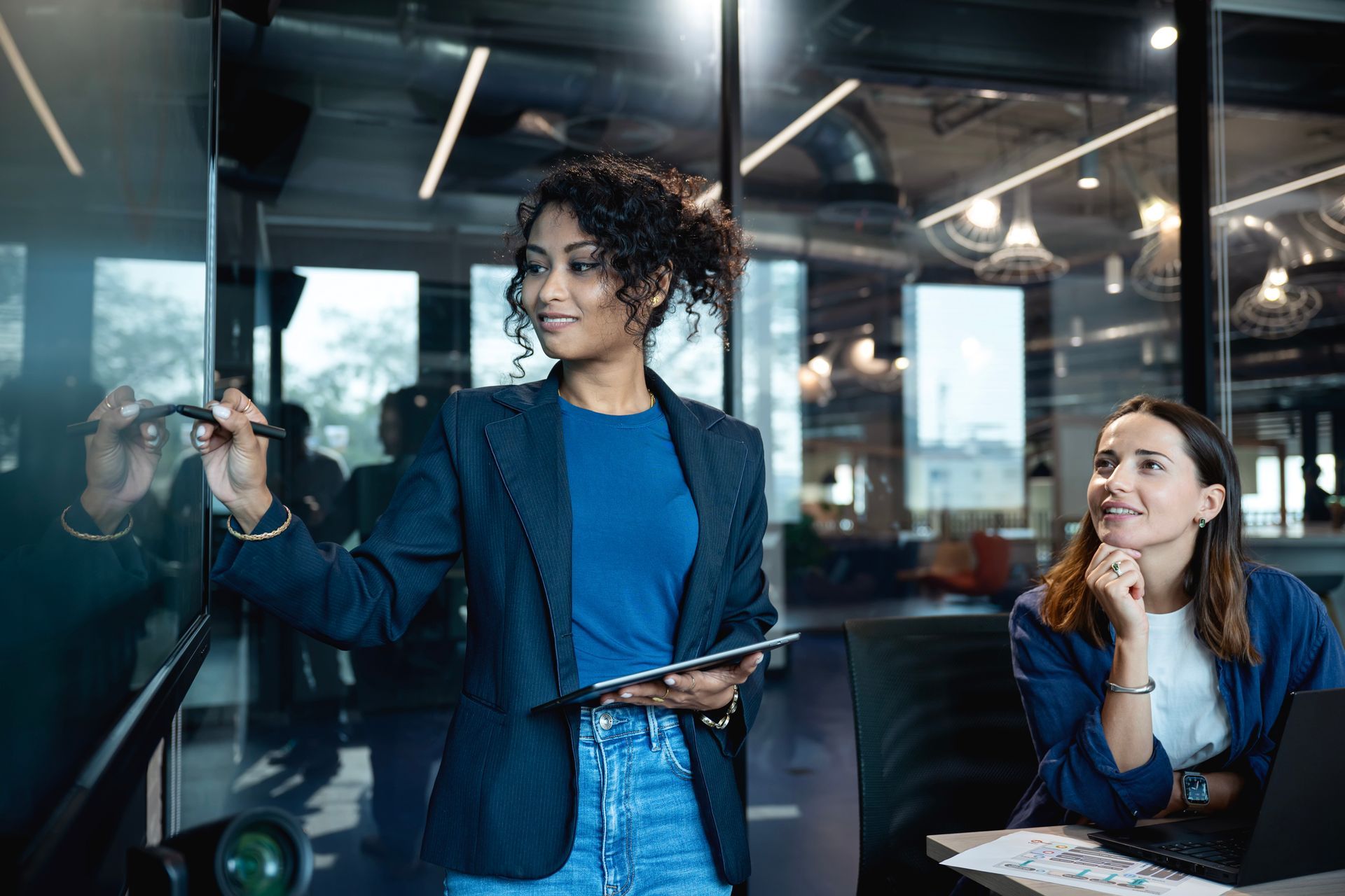 A woman is writing on a whiteboard while another woman looks on.