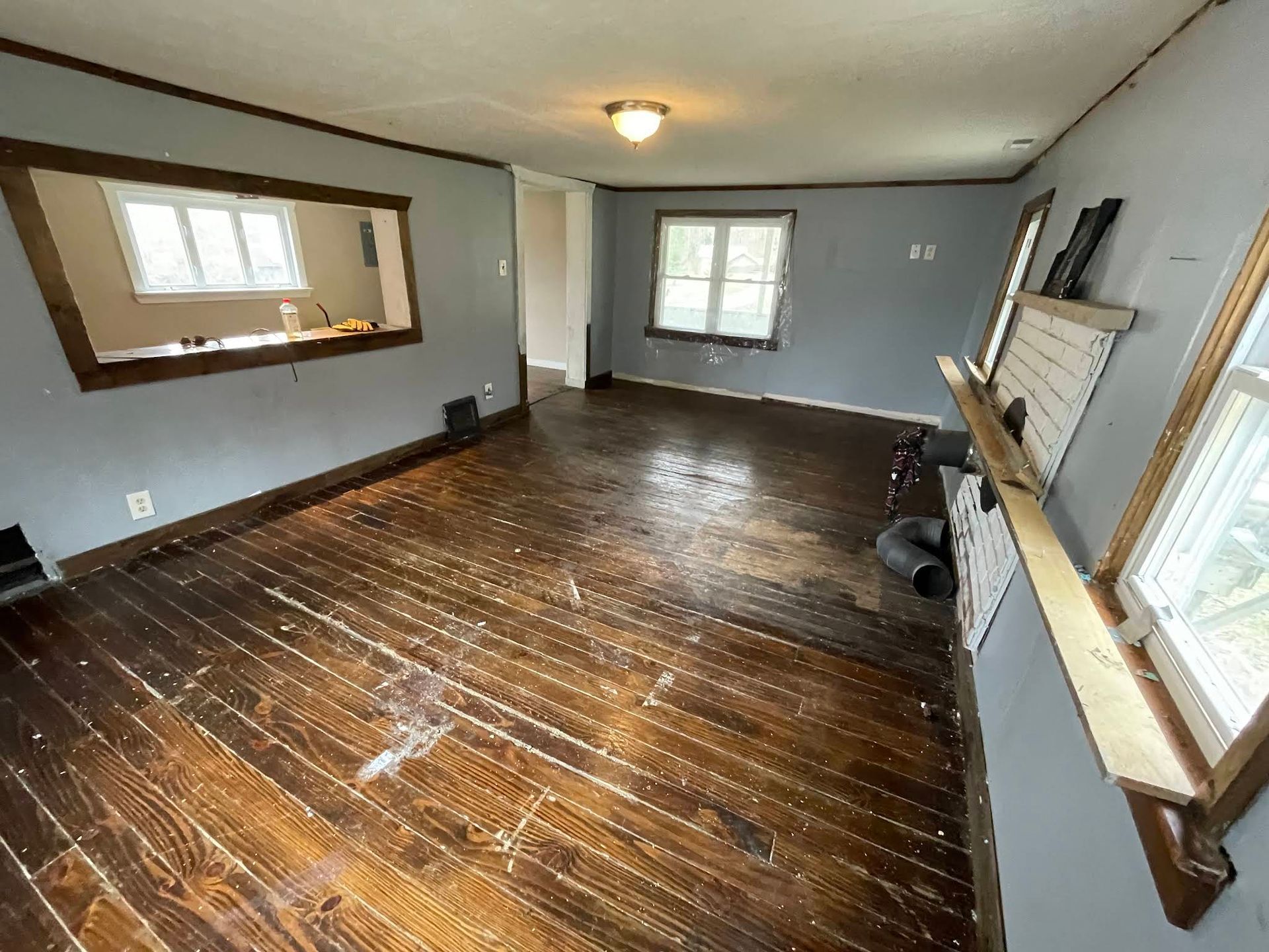 Room with damaged hardwood floors, gray walls, and a pass-through window to the kitchen.
