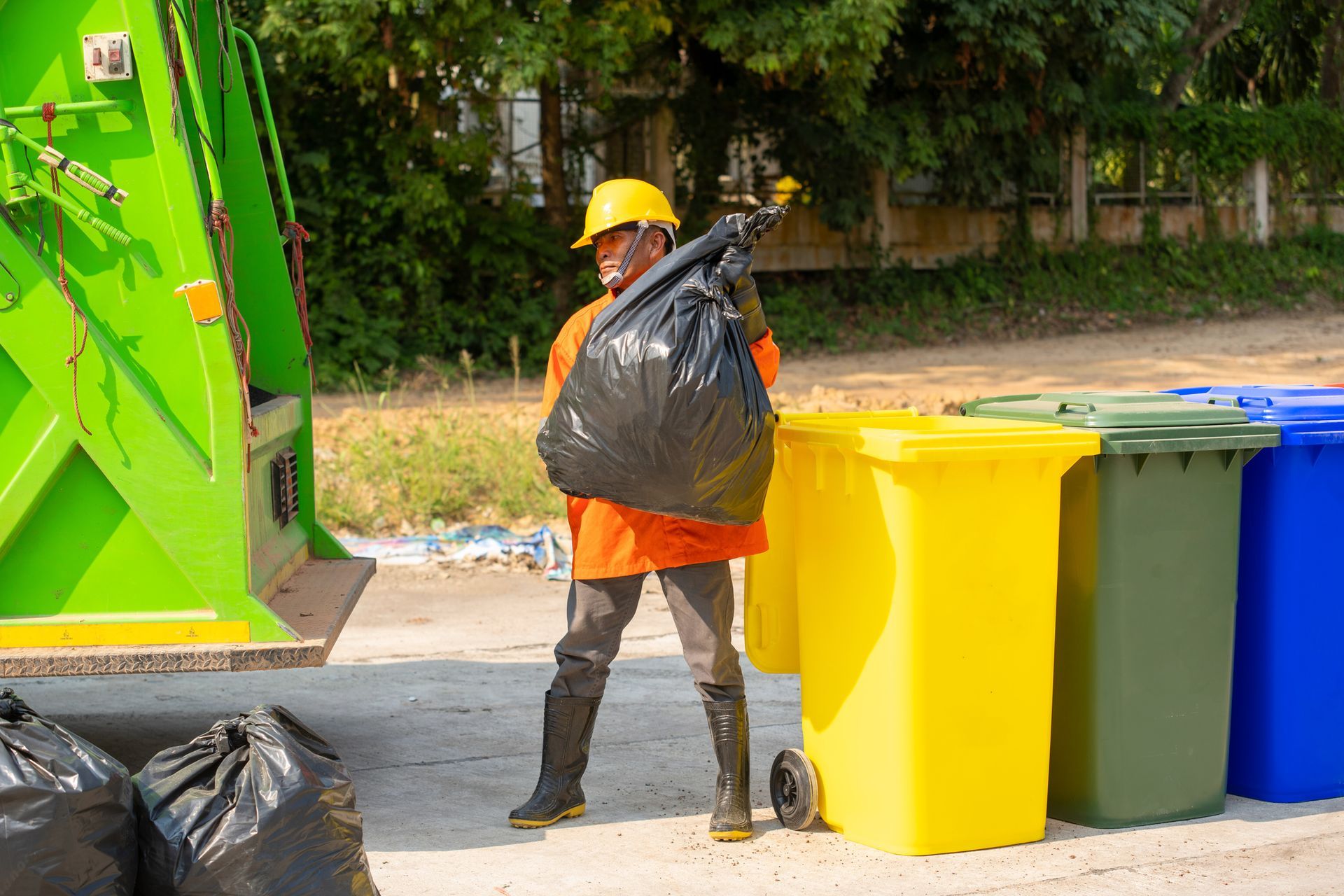 Garbage collector in orange uniform and yellow hard hat carries a black trash bag near recycling bins.