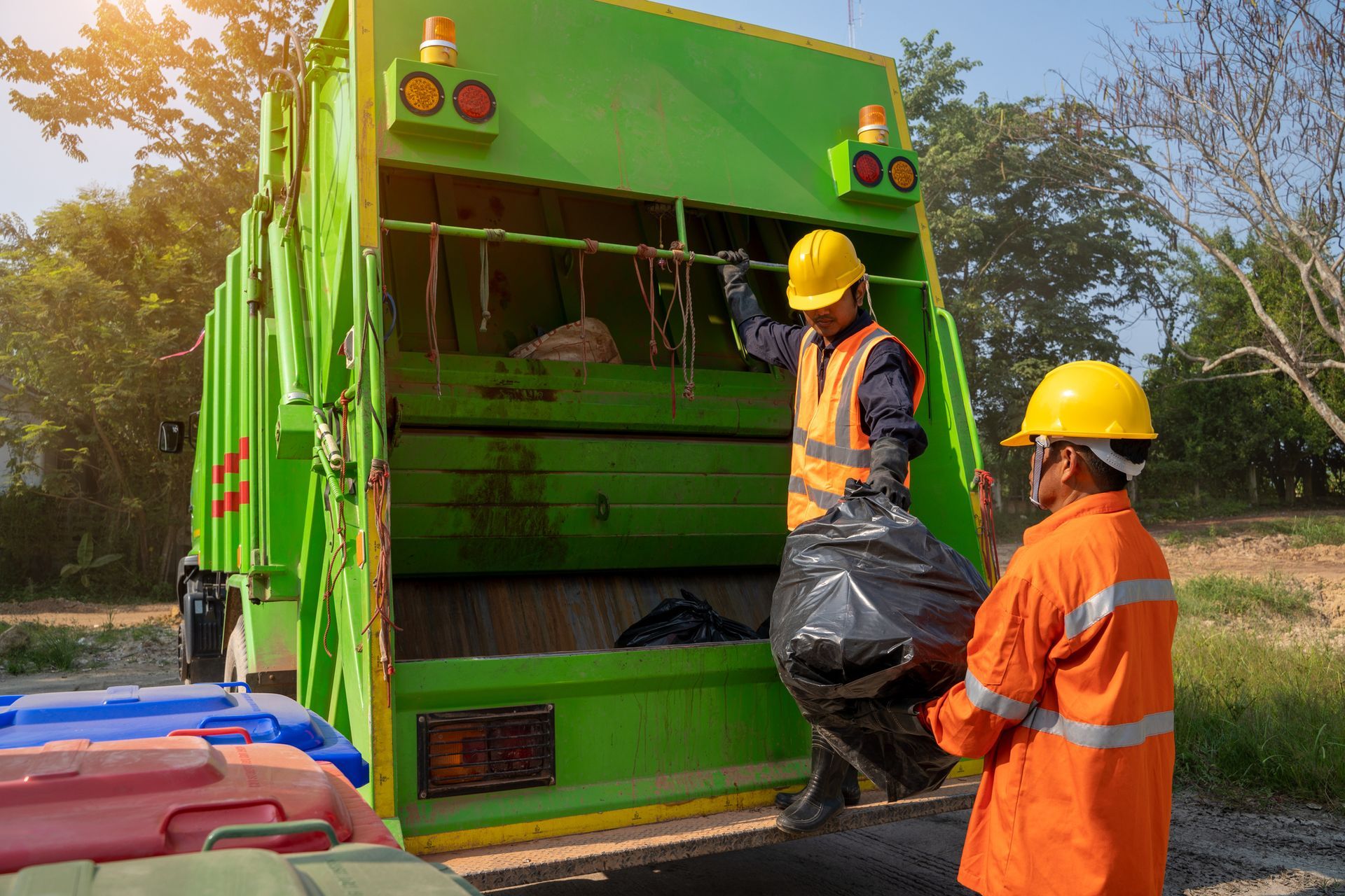 Two sanitation workers in hard hats loading a black trash bag into a green garbage truck.