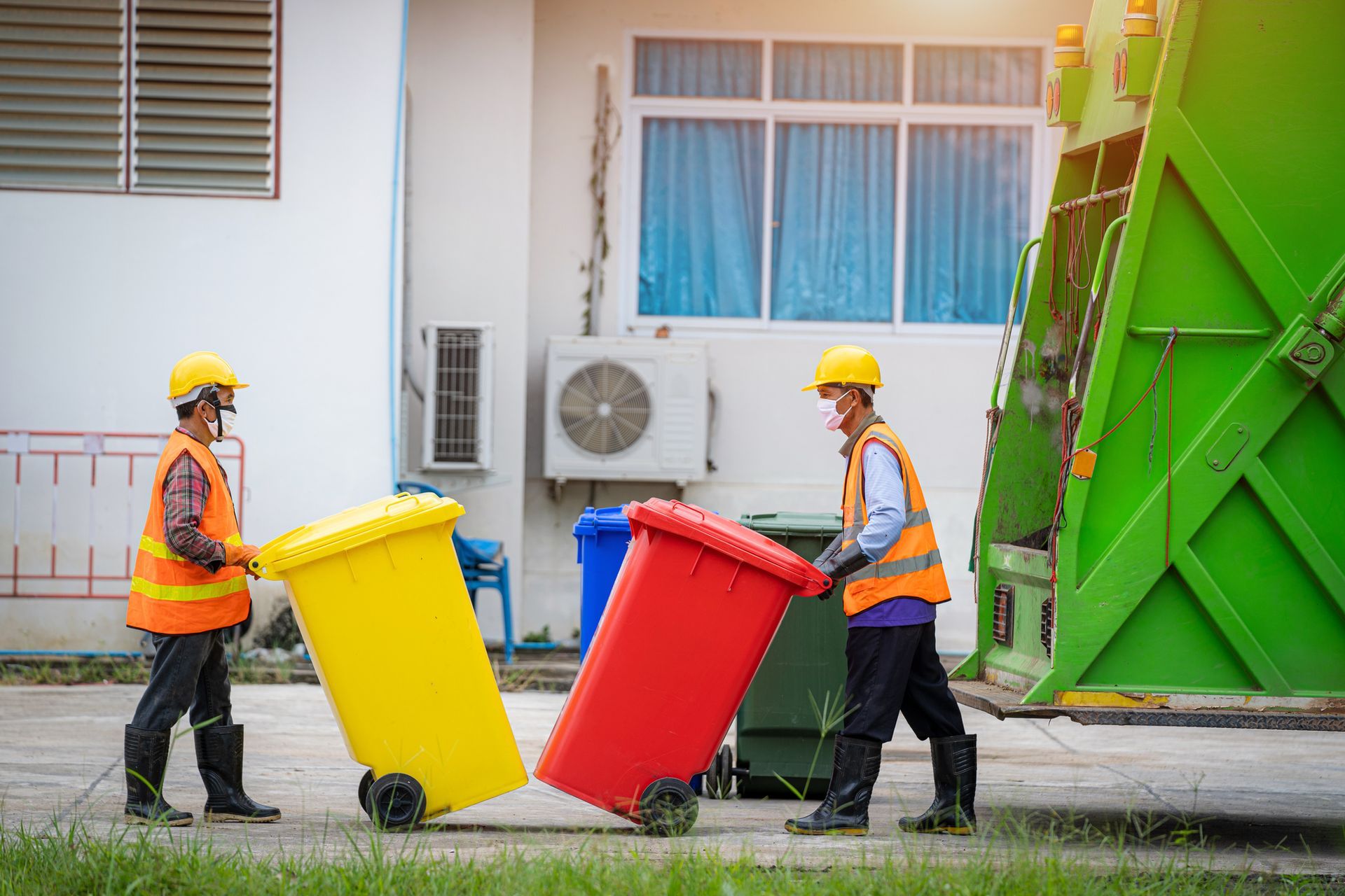 Two sanitation workers in vests and hard hats rolling colorful trash bins toward a garbage truck.