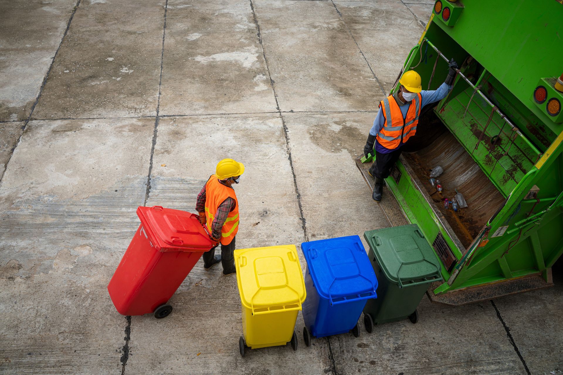 Two sanitation workers loading colorful bins into a garbage truck on a concrete surface.