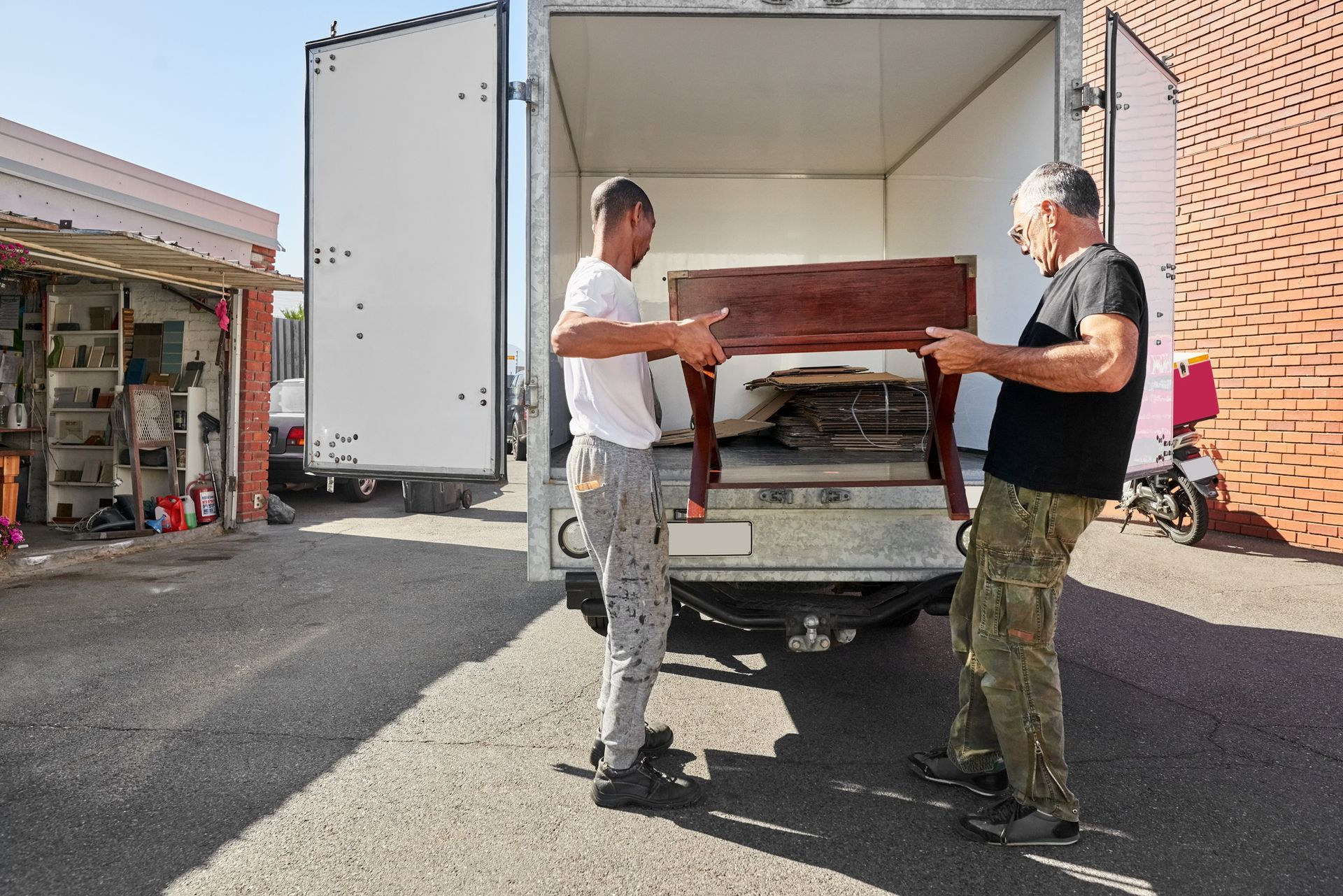 Two people carrying a wooden table toward a moving truck, outdoors.