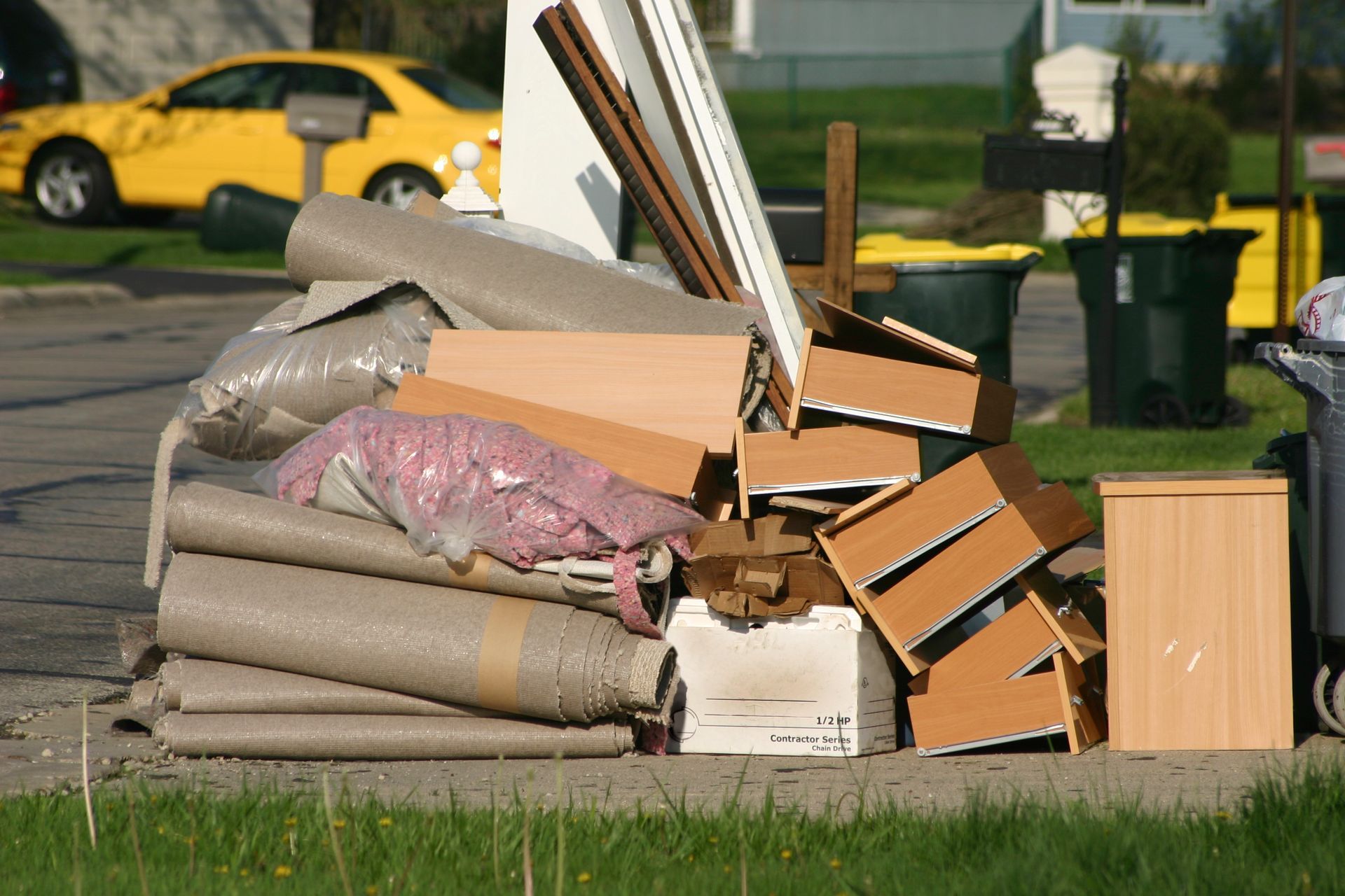 Pile of discarded household items, including cardboard boxes and rolled-up carpet, on a residential street.