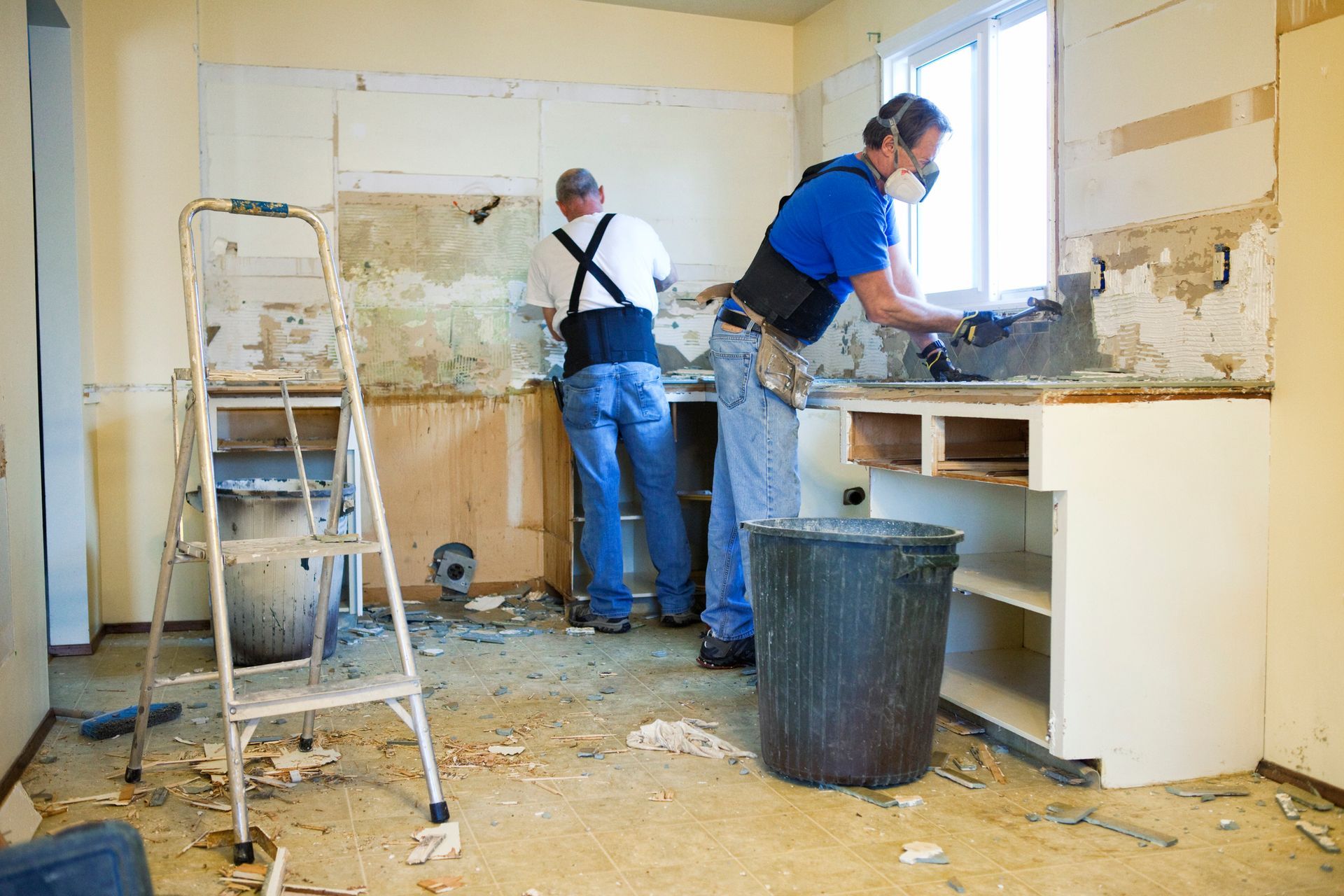 Two people renovating a kitchen, removing tiles and drywall. A ladder, trash can, and debris are present.