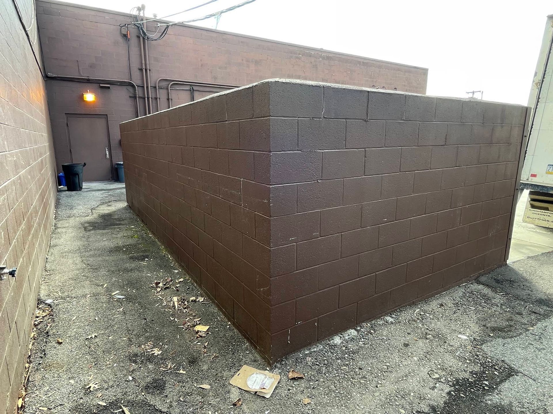 Brown cinder block structure in an alley, against a brown building with a door.