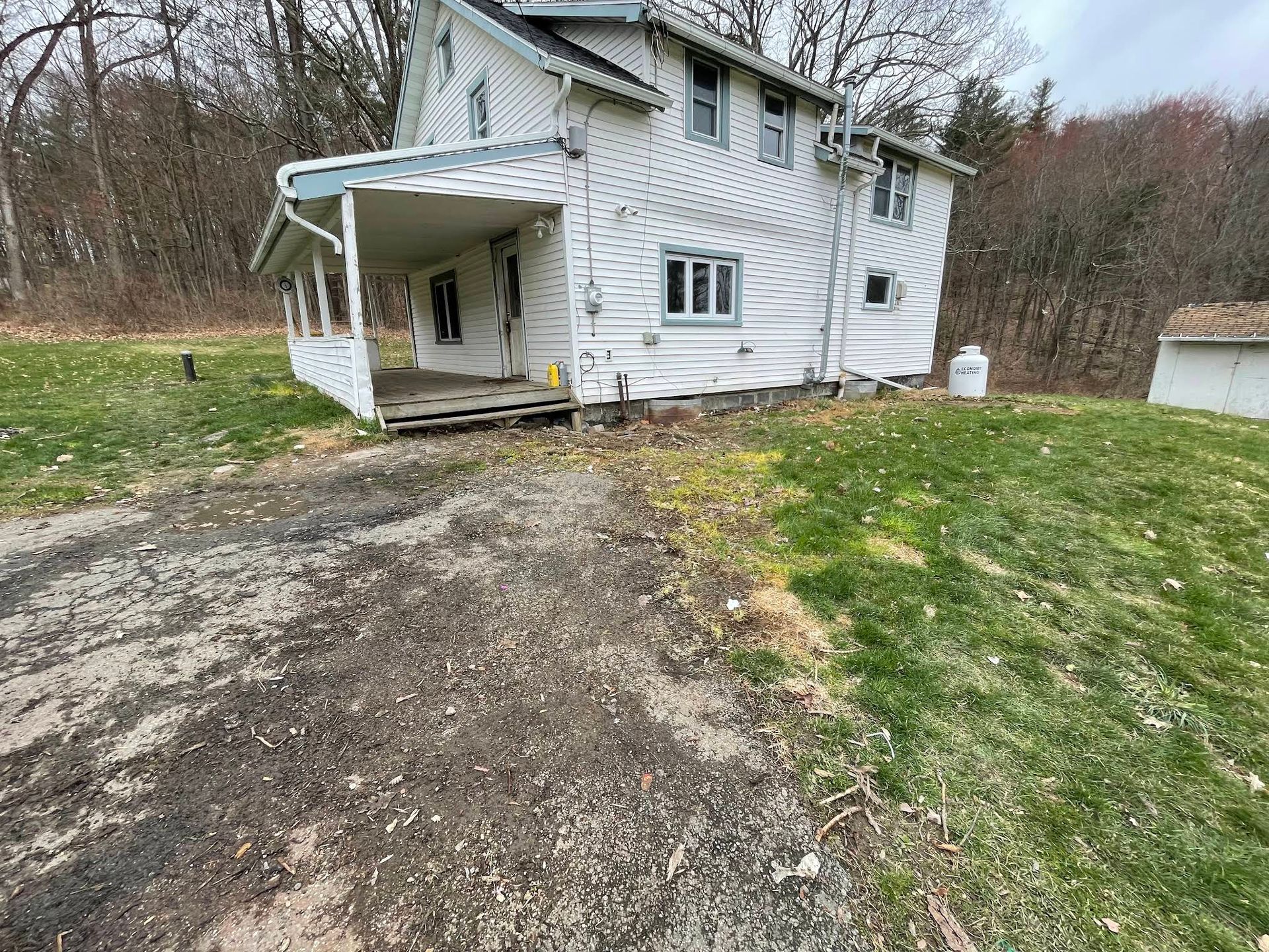 Dilapidated white house with porch, gravel driveway, and surrounding grass, on a cloudy day.