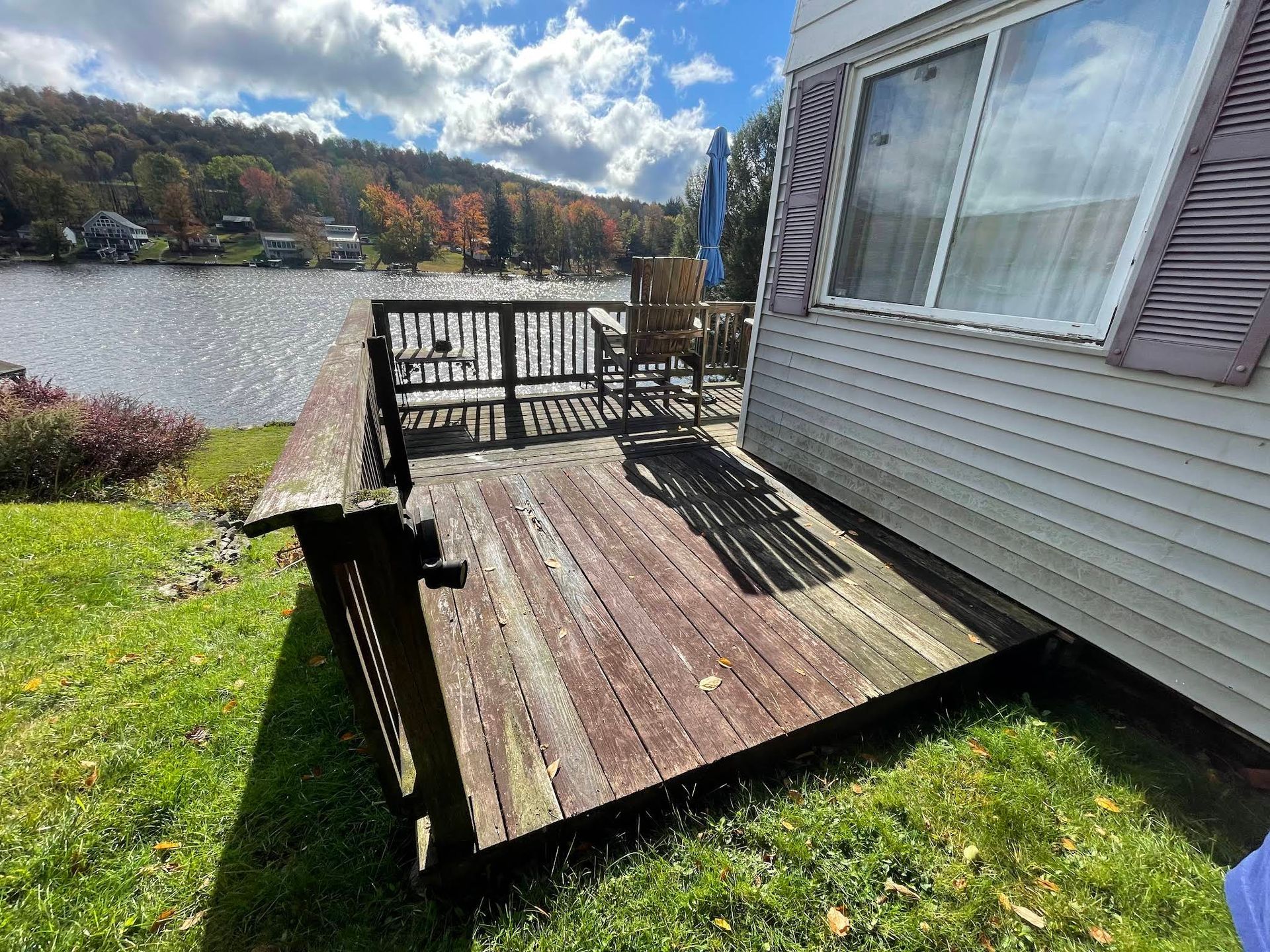 Wooden deck next to a lake. House siding on right, green grass, and trees in the background.