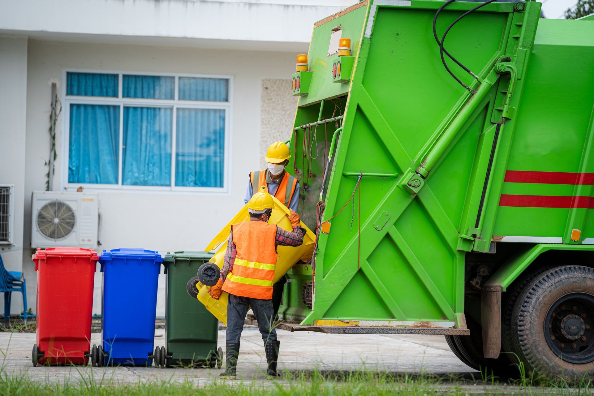 Two sanitation workers emptying a yellow bin into a green garbage truck. Three colored bins in front.