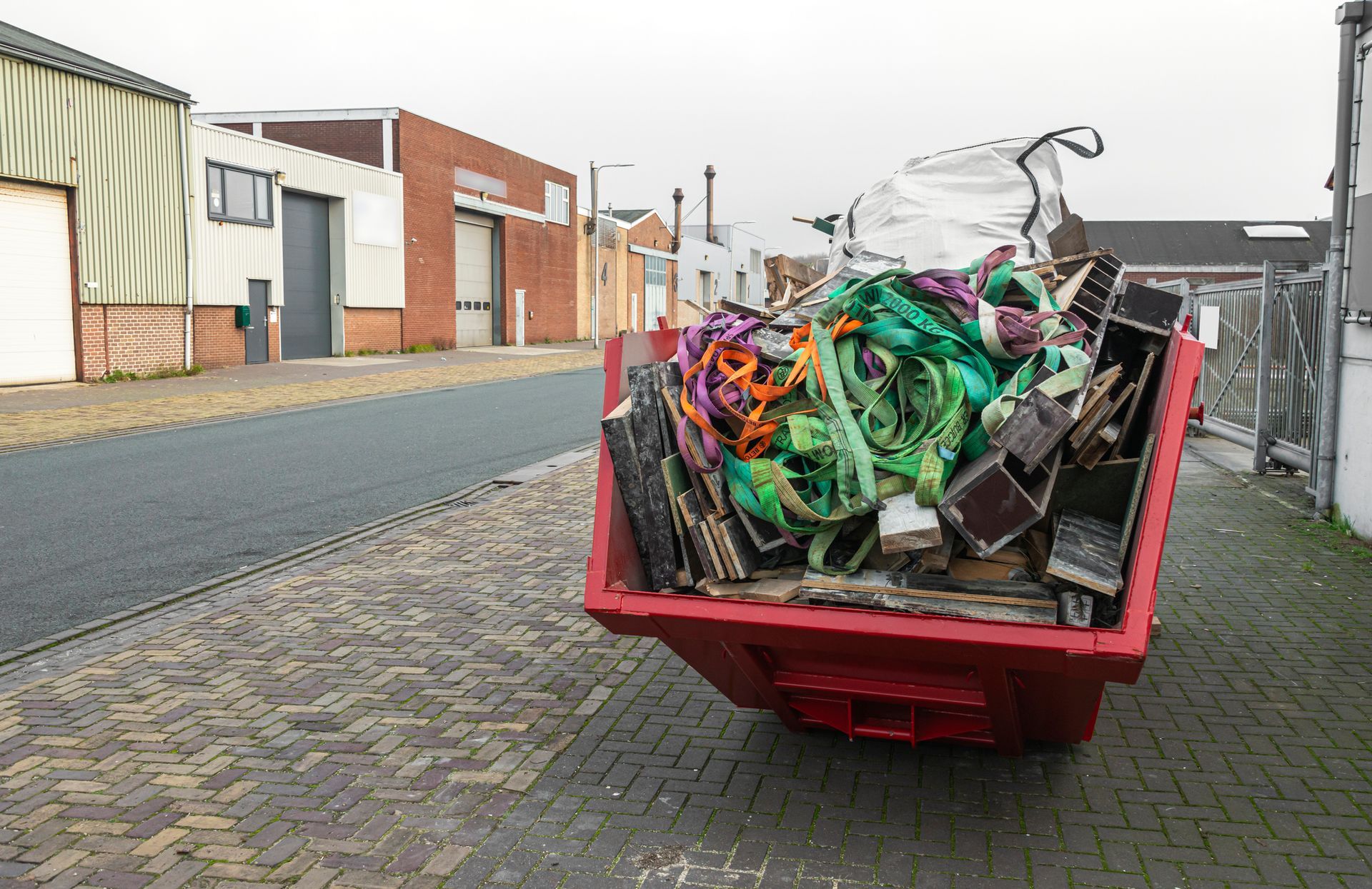 Red dumpster overflowing with trash on a brick-paved street, warehouses in background.