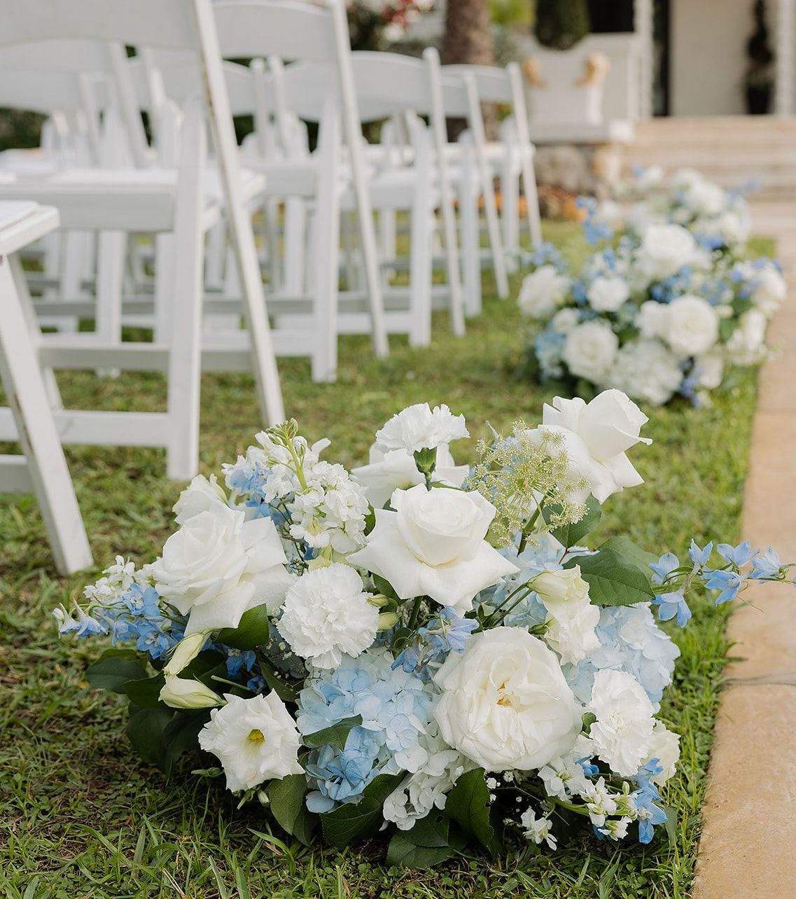 White chairs line a green lawn, with flower arrangements of white and blue blooms marking the aisle for an outdoor wedding.
