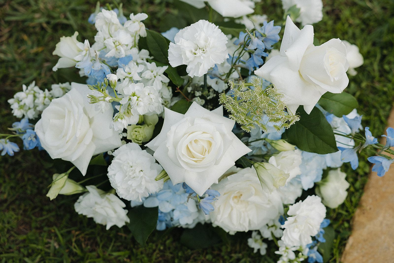 Close-up of a floral arrangement featuring white roses, blue delphiniums, and white carnations against a green lawn.