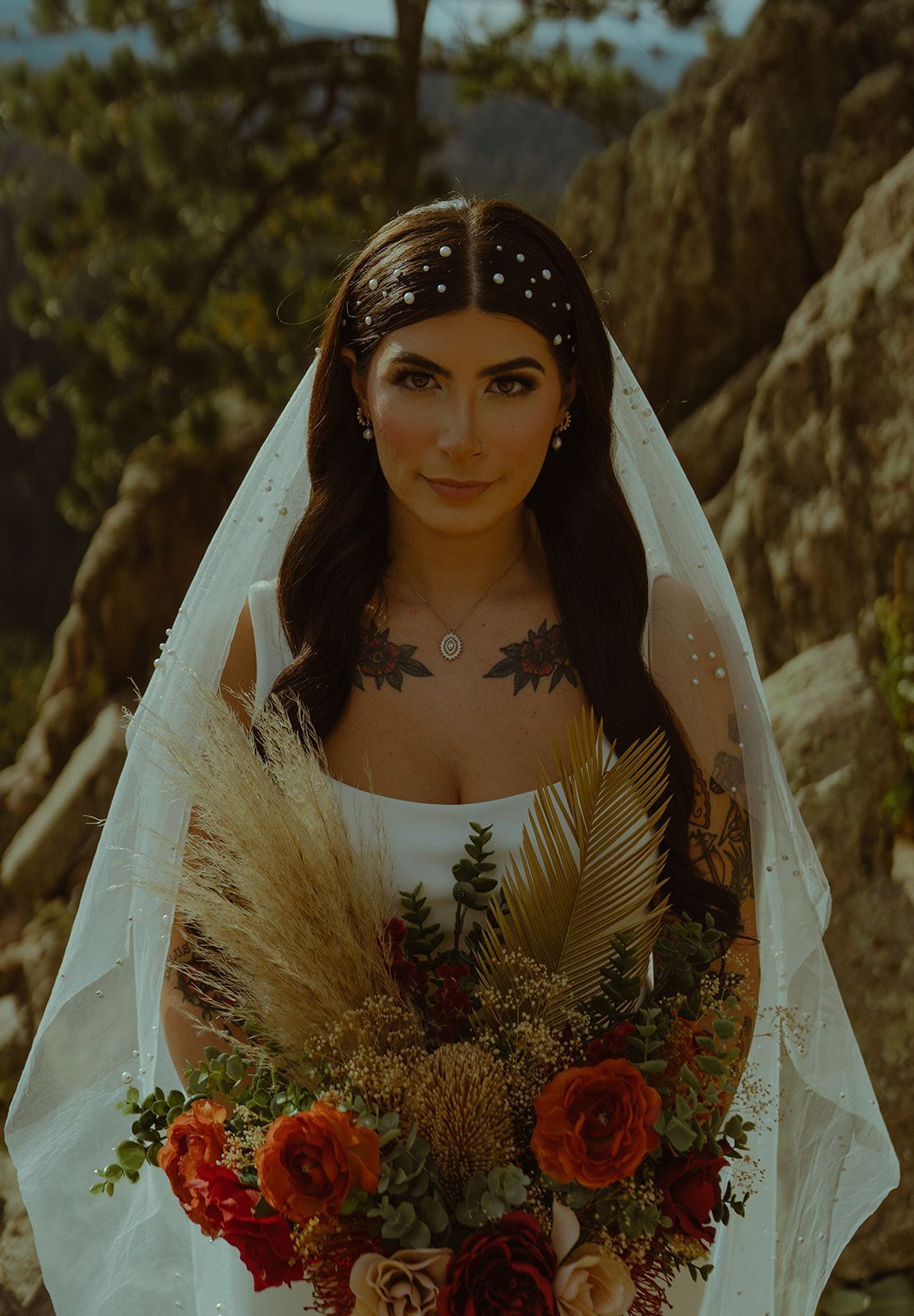Bride in a white dress and veil holding a bouquet of flowers, standing outdoors with rocks in the background.