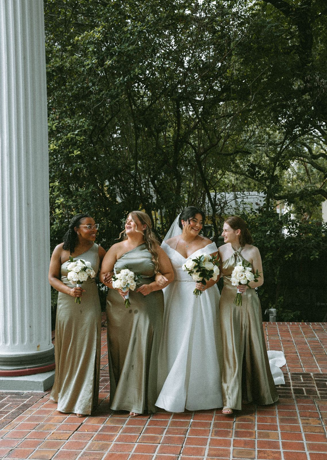 Bride and bridesmaids in long, olive green dresses pose outdoors by a column. They smile and hold bouquets of white flowers.