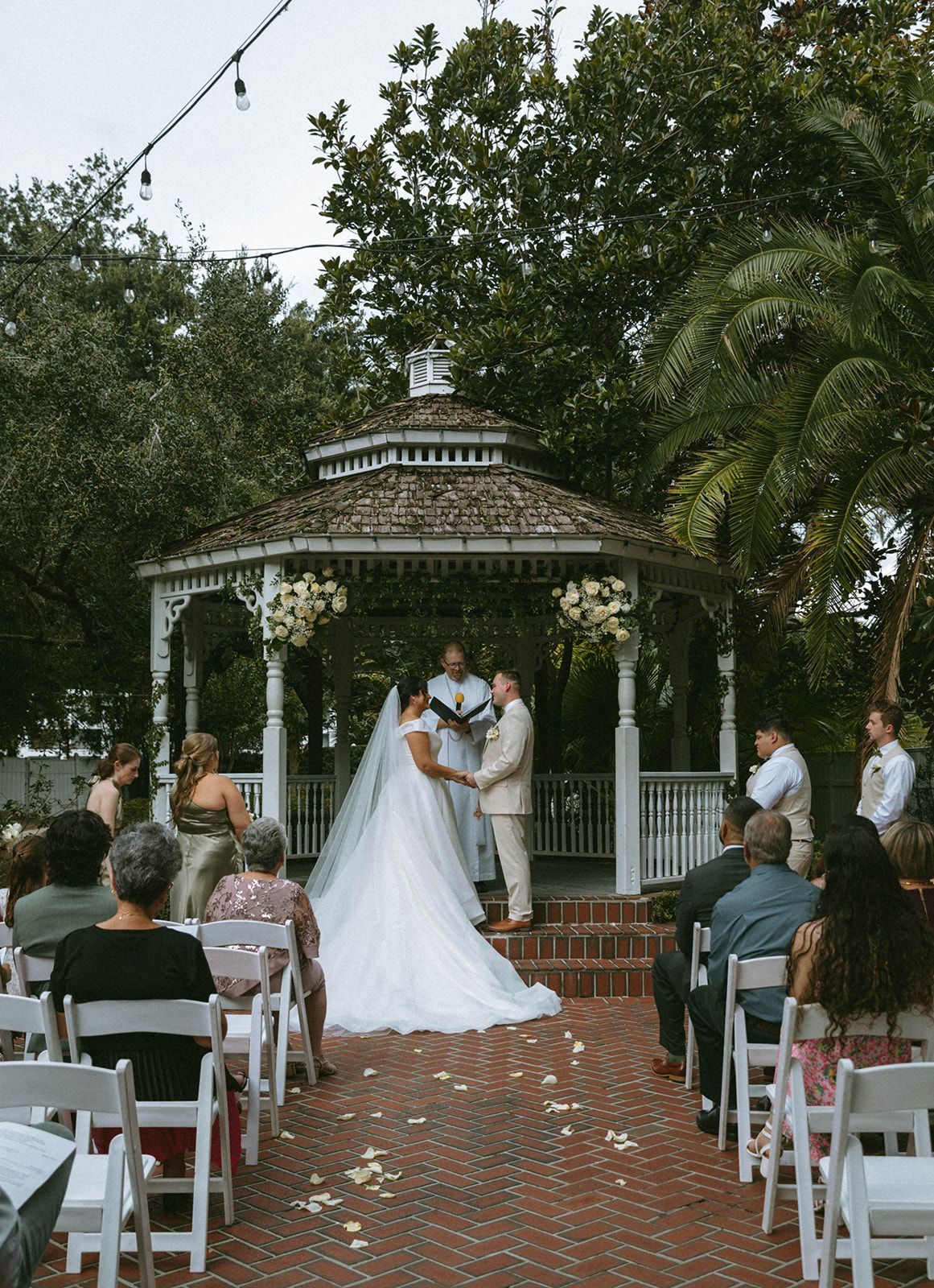 Wedding ceremony in a garden setting. Bride and groom stand under a gazebo, exchanging vows, with guests seated in white chairs.