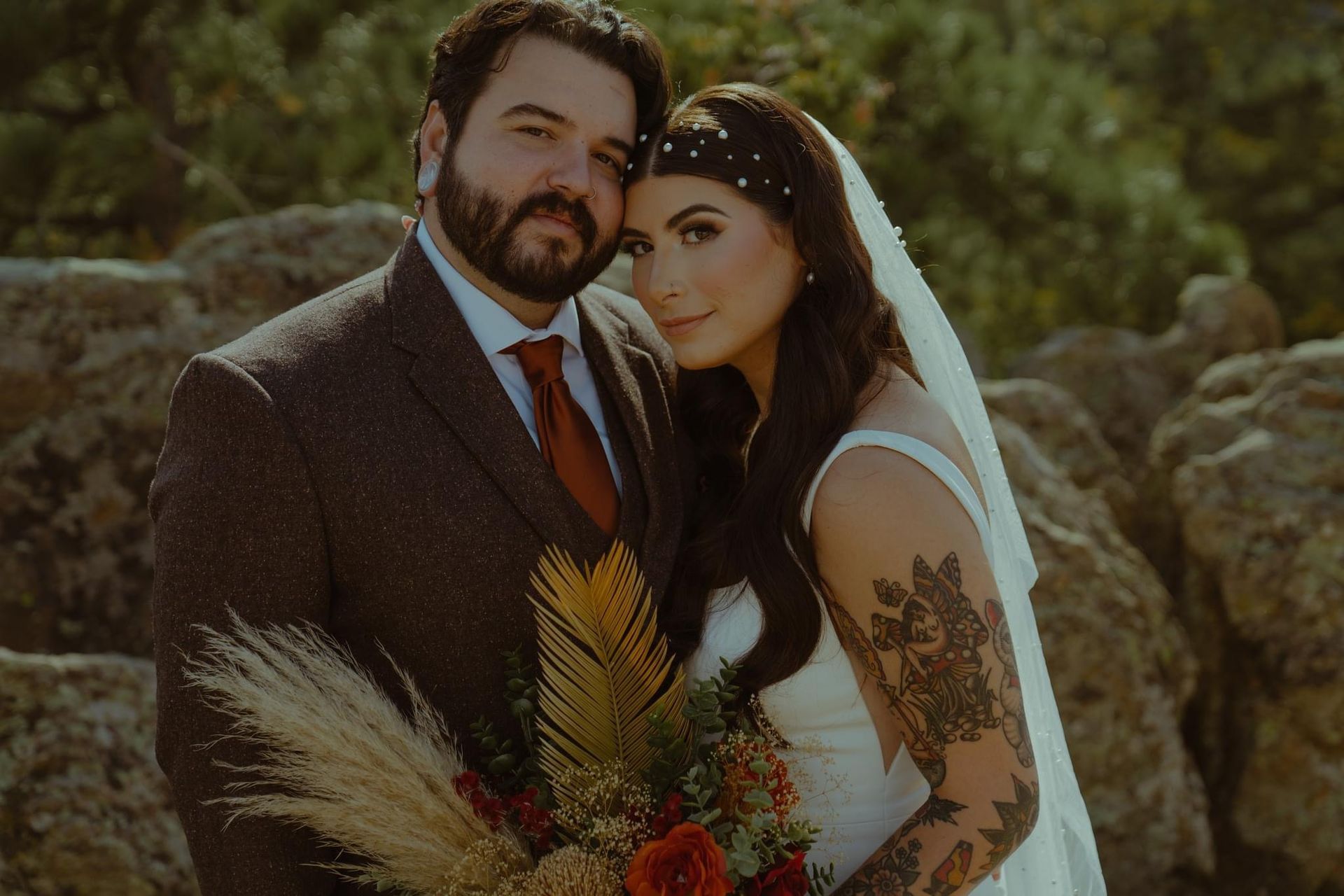 Newlyweds, a man and woman, pose together outdoors. The woman wears a wedding dress and veil, and the man wears a suit. They are near some rocks.