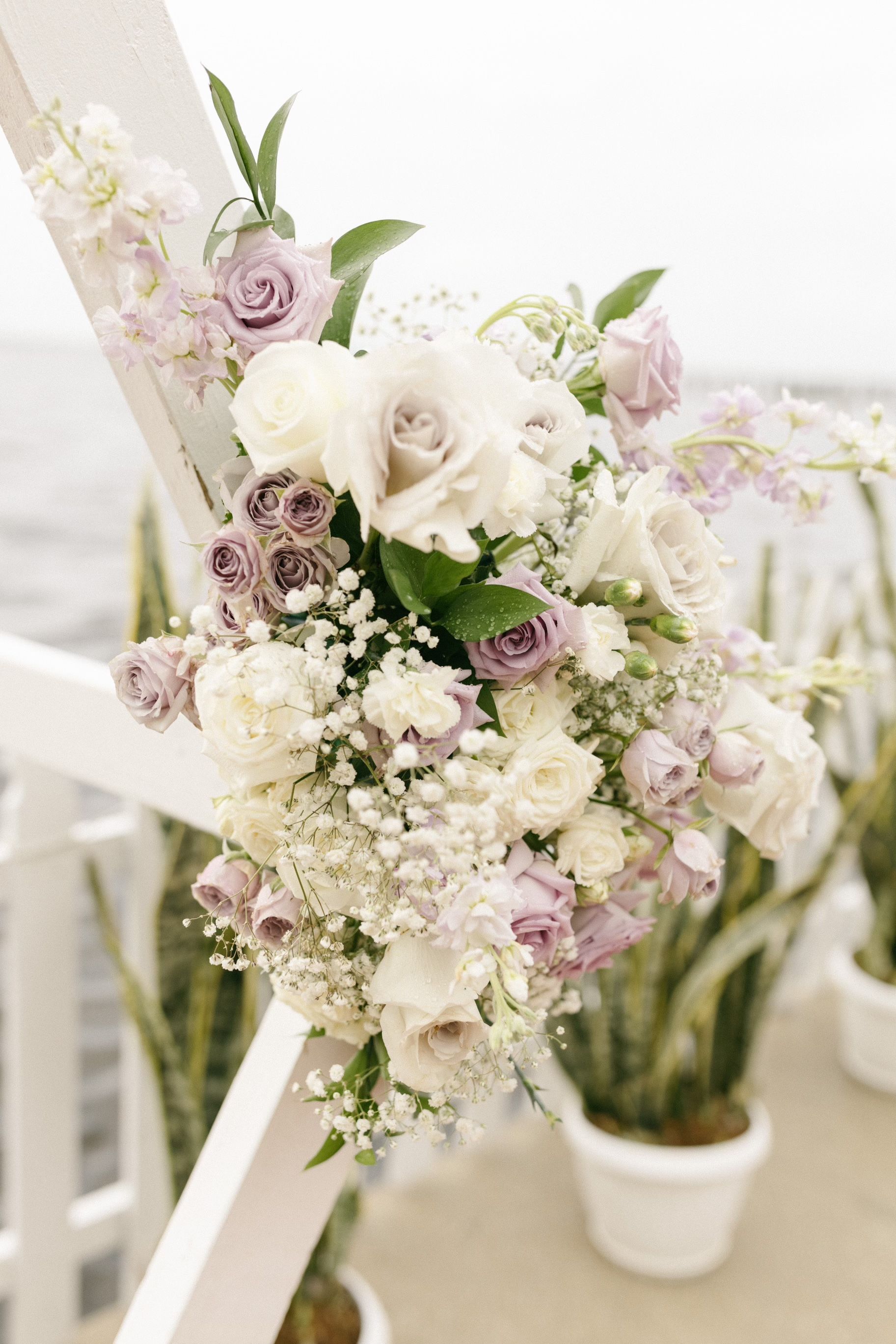 Wedding arch decorated with white, lavender, and purple roses, with baby's breath, against a blurred ocean backdrop.