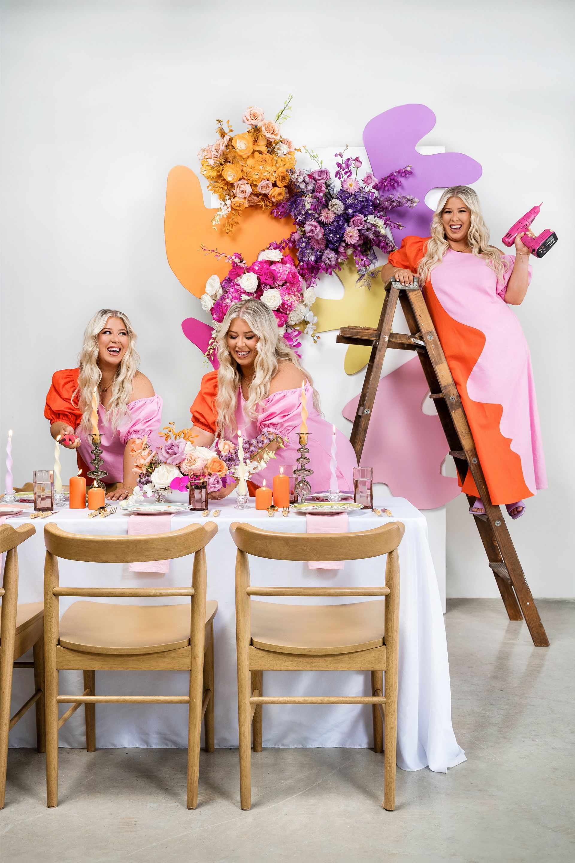 Three blonde women in colorful dresses at a decorated table, one on a ladder with a drill. Colorful abstract floral backdrop.