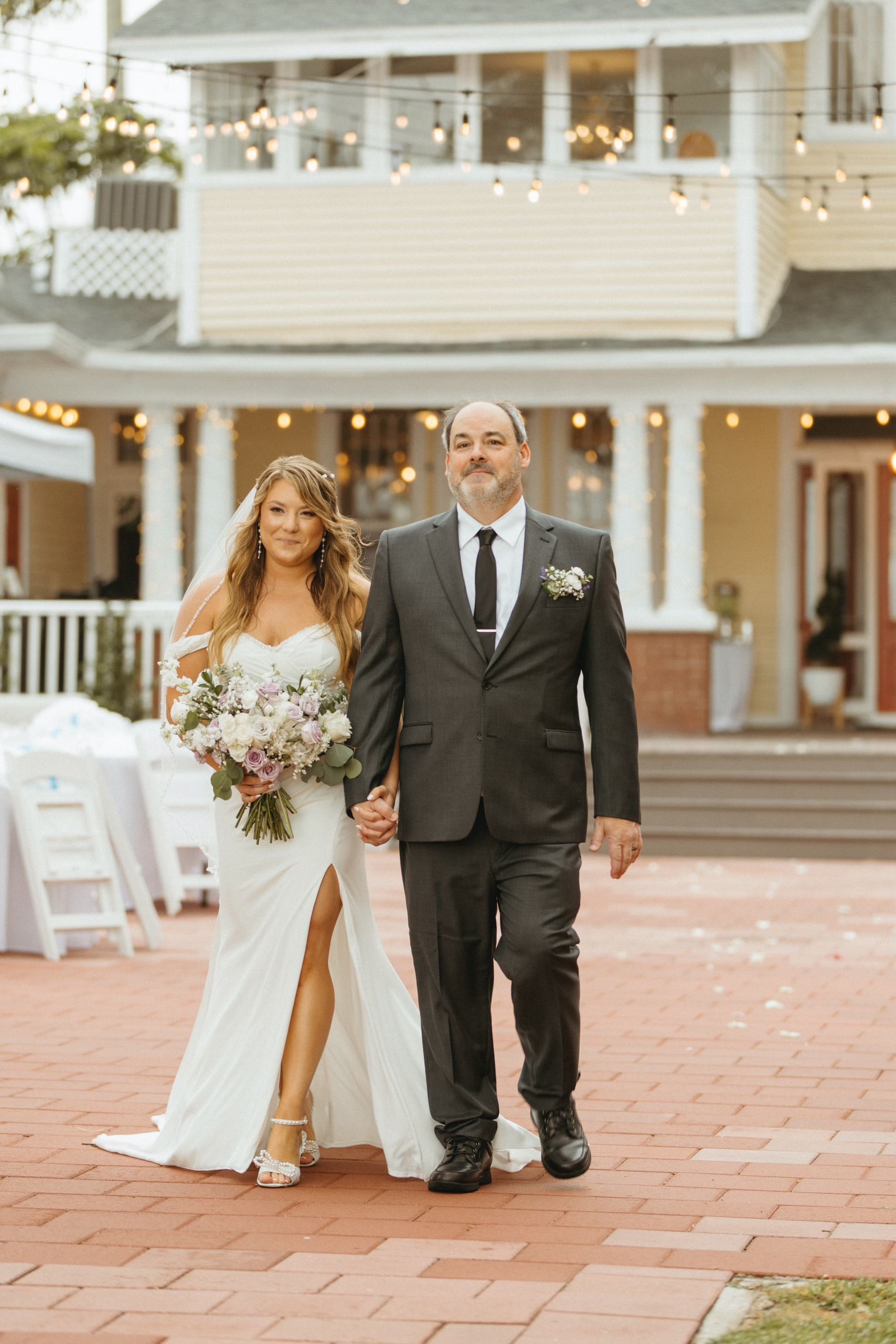 Bride in white gown, holding hands with a man in a suit, walking on a brick path towards a yellow house with string lights.