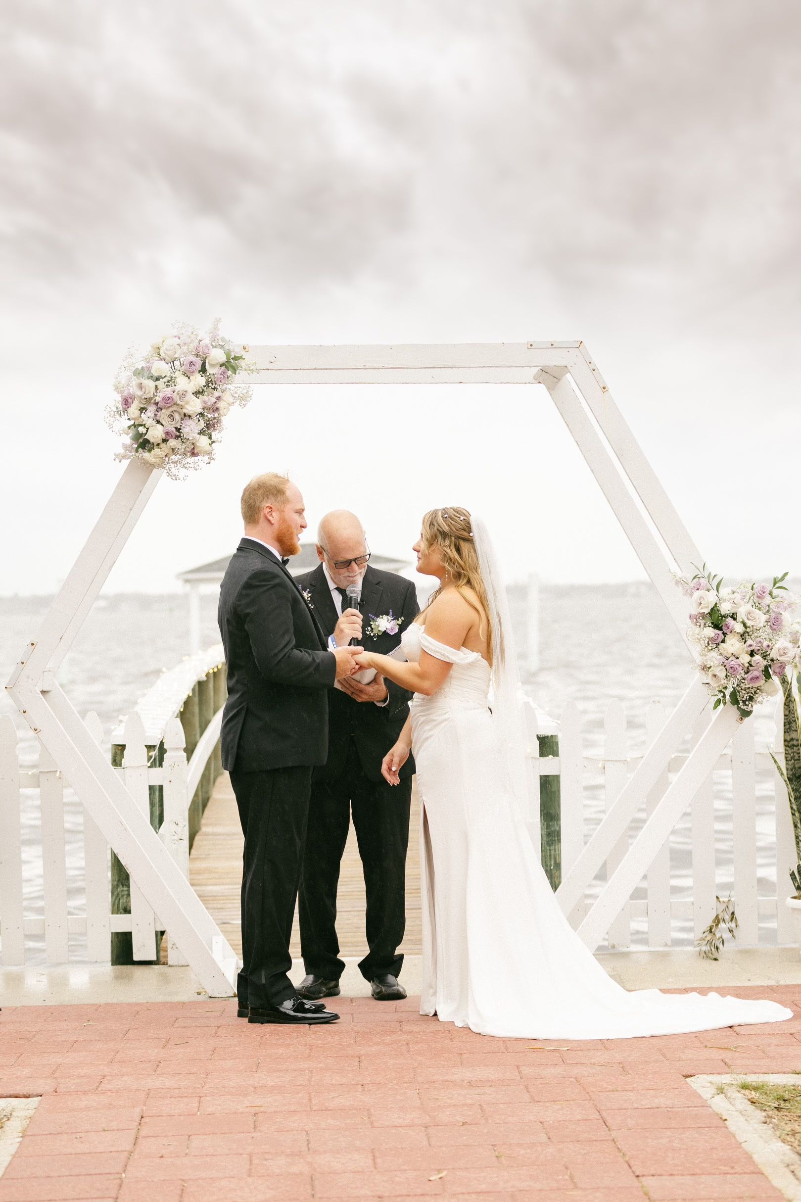 A bride and groom exchange vows under a white hexagonal archway on a pier. The officiant stands between them.