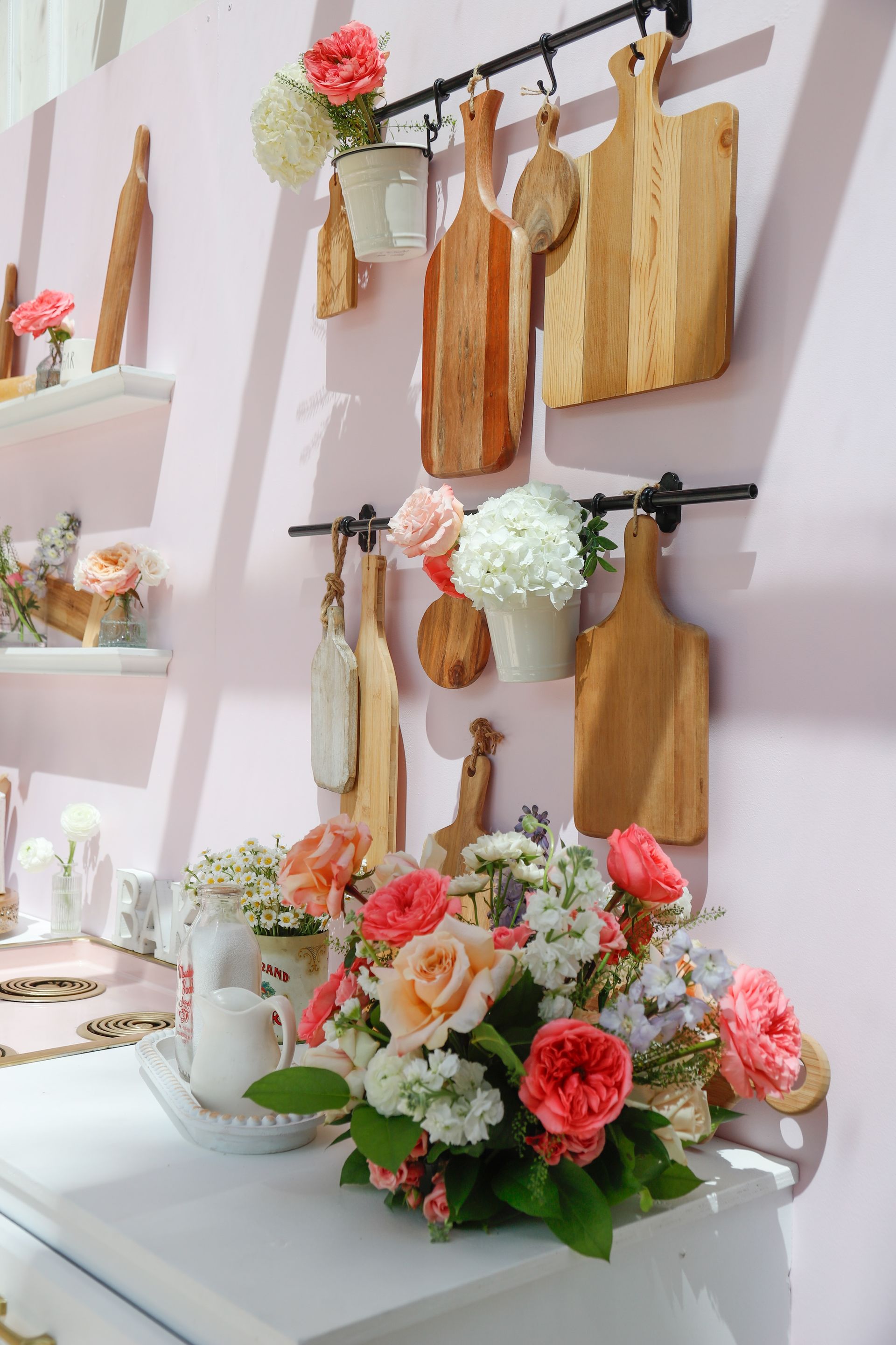 A pink kitchen scene featuring wooden cutting boards, flowers, and decorative shelves.