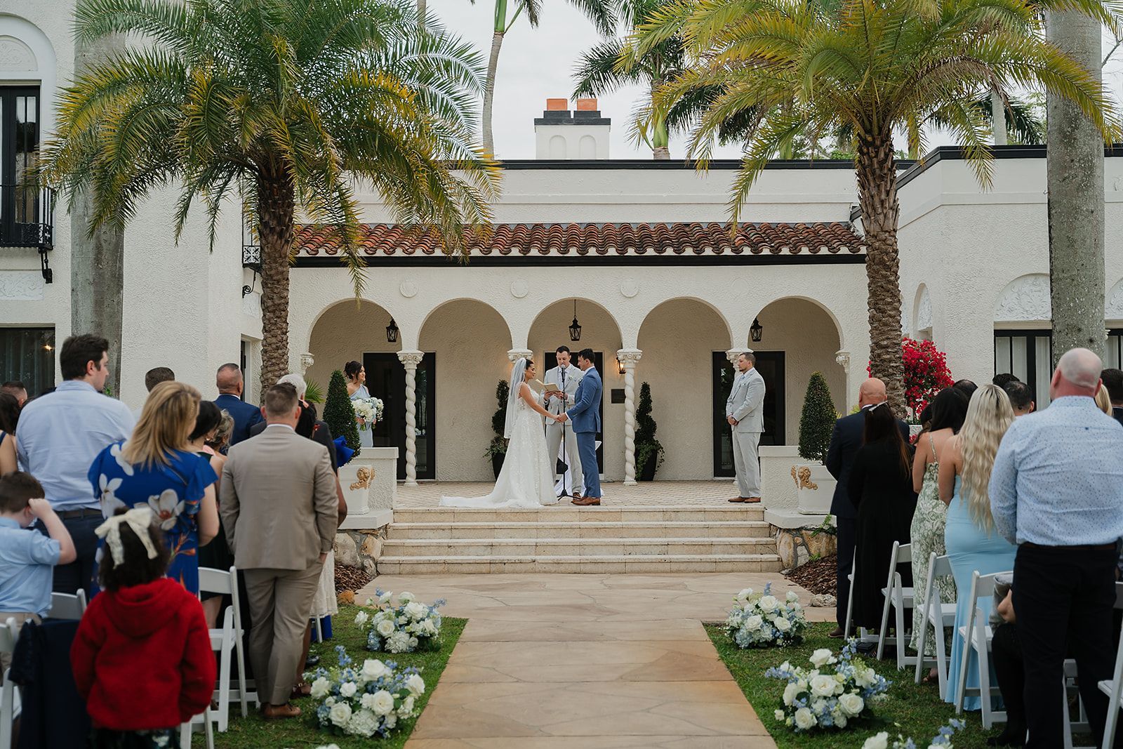Wedding ceremony outside a Spanish-style building. A couple stands at an altar as guests watch. Palm trees frame the building.