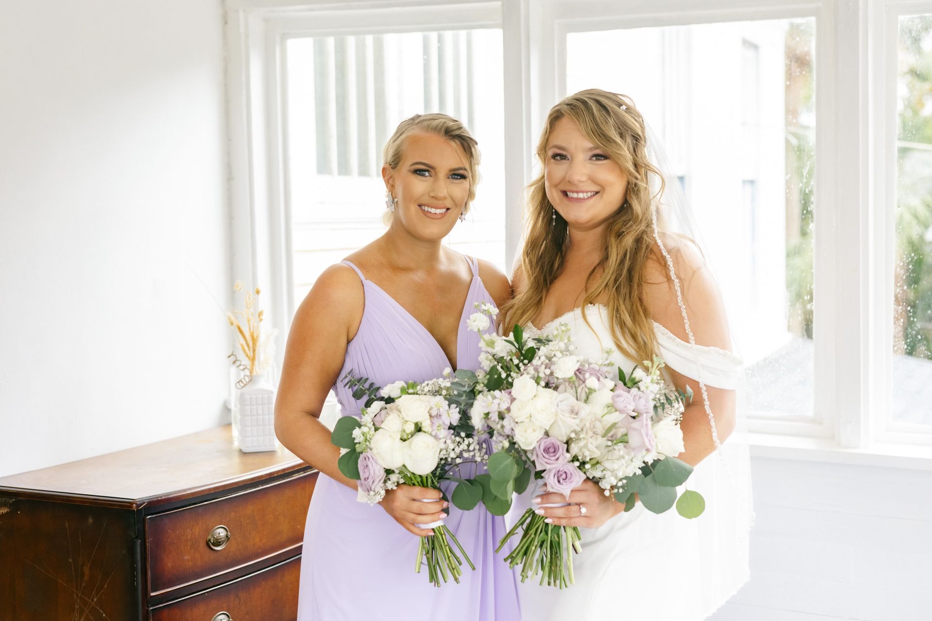 Bride and bridesmaid smiling, holding bouquets. Bride in white gown, bridesmaid in lilac dress, standing by a window.