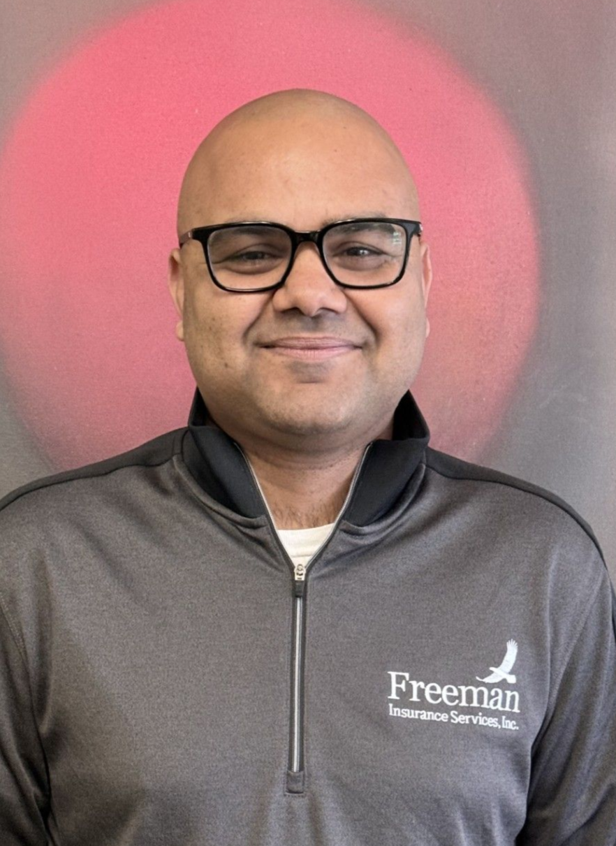 Man with glasses smiles, wearing a gray Freeman Services zip-up jacket in front of a red backdrop.