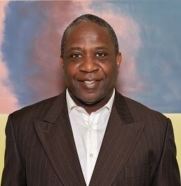 Man in brown suit and white shirt, smiling, in front of colorful wall.