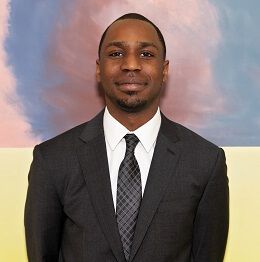 Man in a suit smiles, standing indoors in front of a colorful abstract painting.