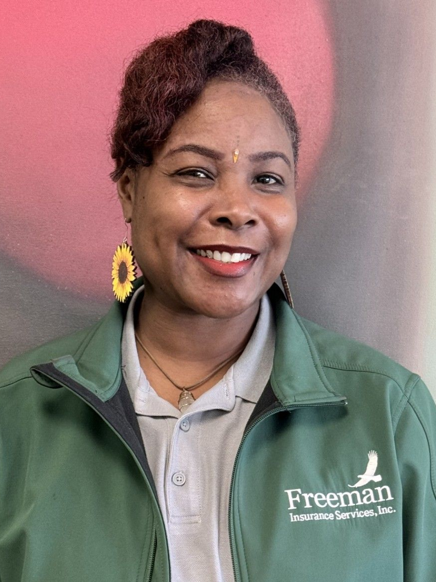 Woman in green jacket smiling, wearing sunflower earrings, standing in front of a colorful backdrop.