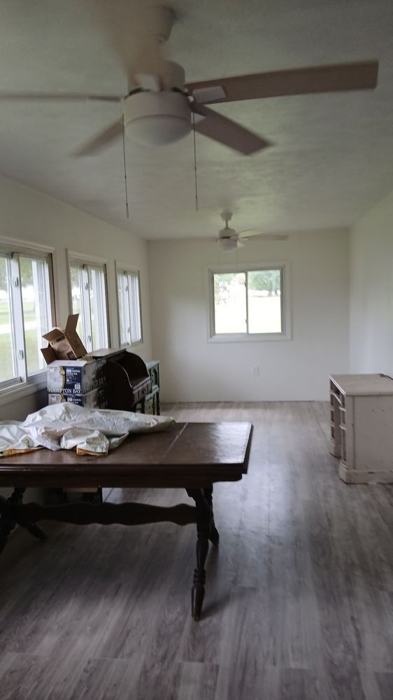 Bright empty living room with ceiling fans, windows, table, and a person sitting on the left