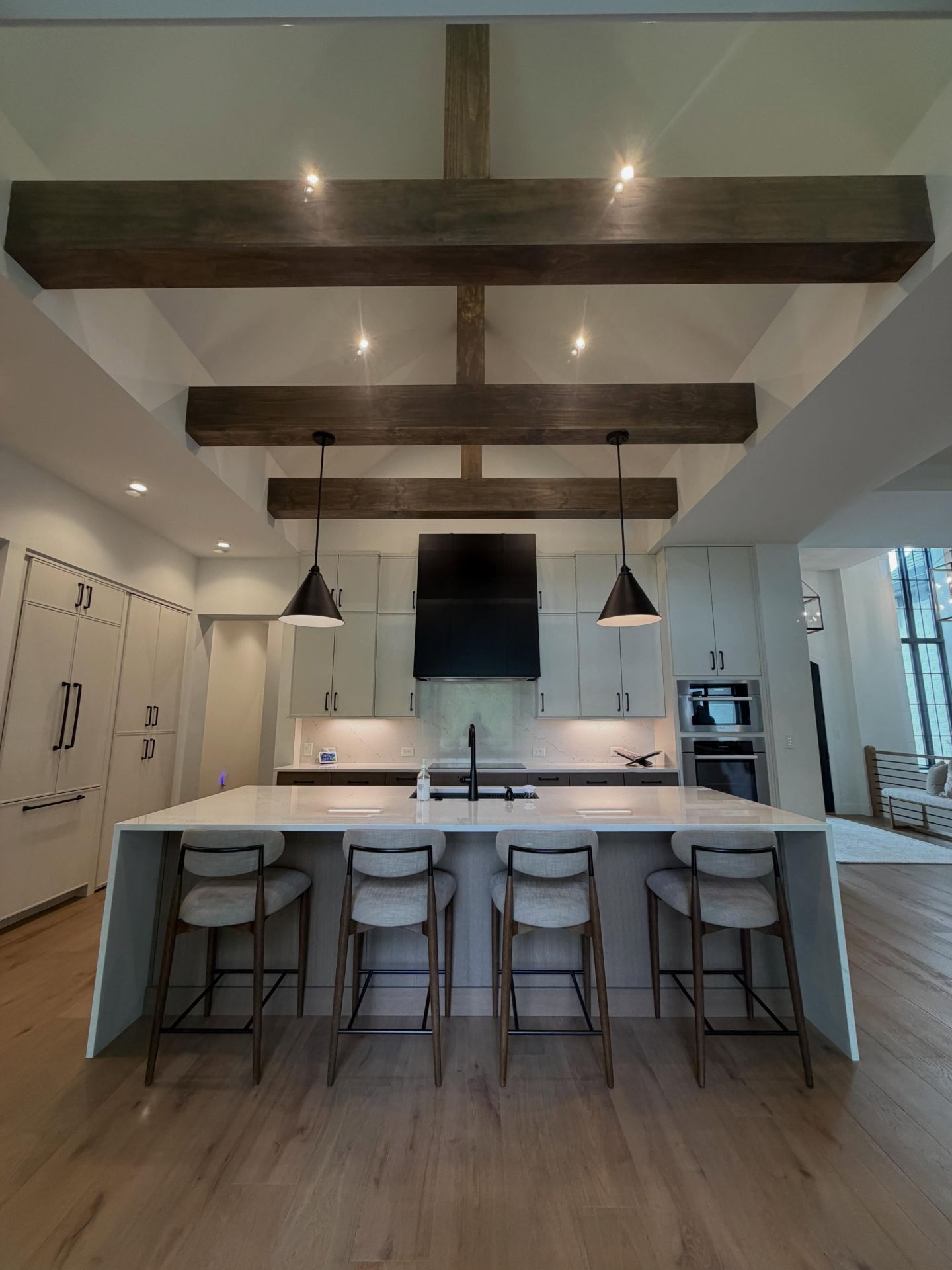 Modern kitchen with white island, bar stools, pendant lights, and dark wood ceiling beams.