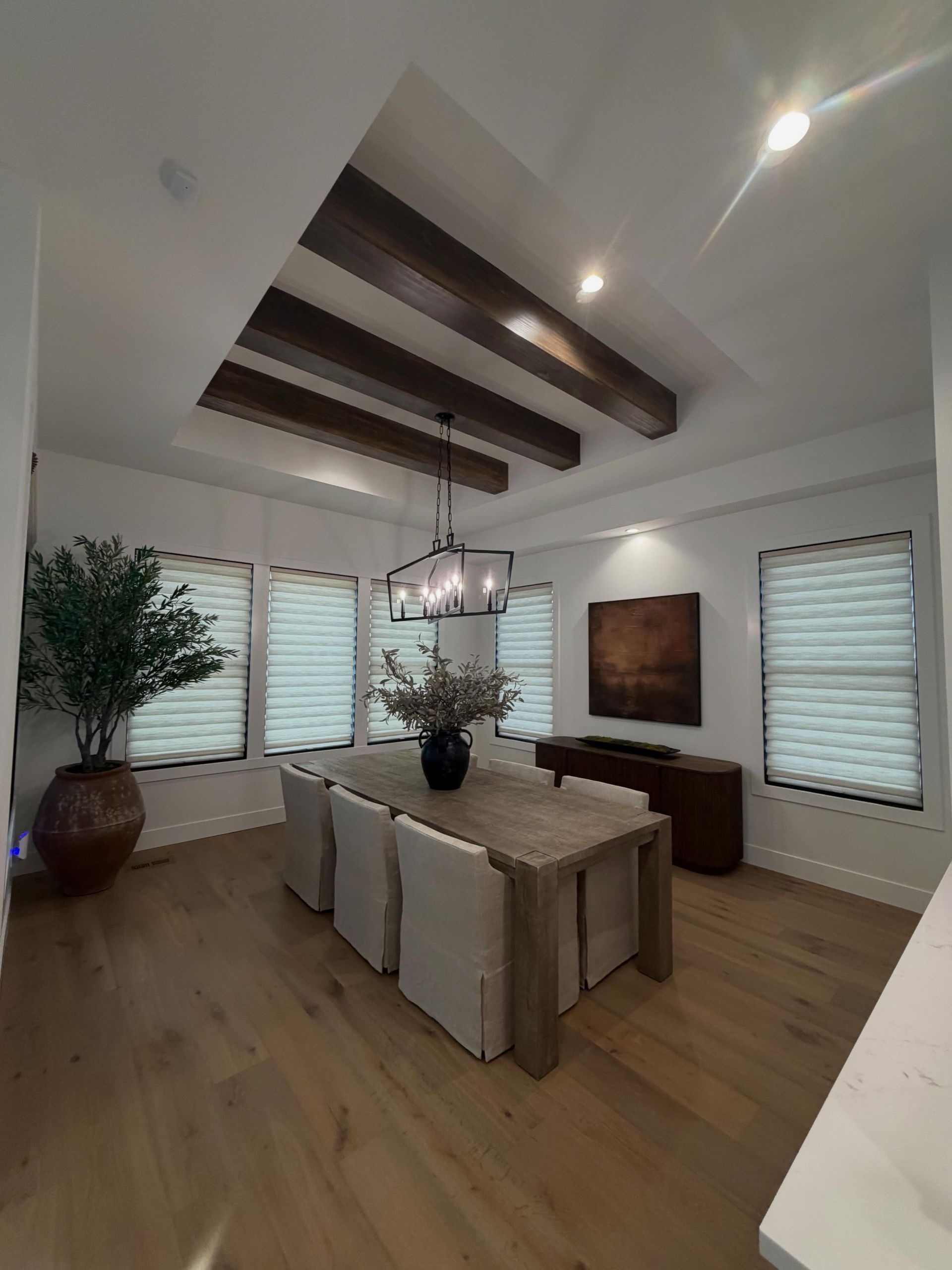 Modern dining room with a long table, beige chairs, wood beams, potted plants, and large windows with blinds