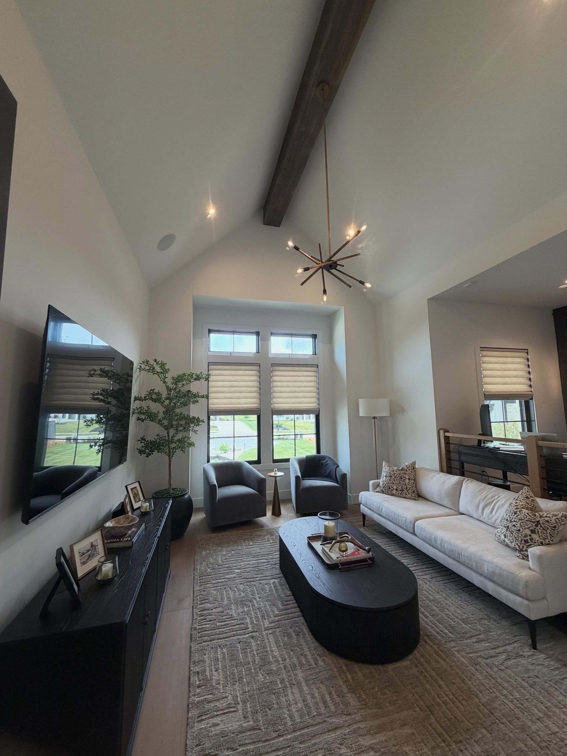 Modern living room with vaulted ceiling, gray sofa, black chairs, wall TV, coffee table, and large windows.