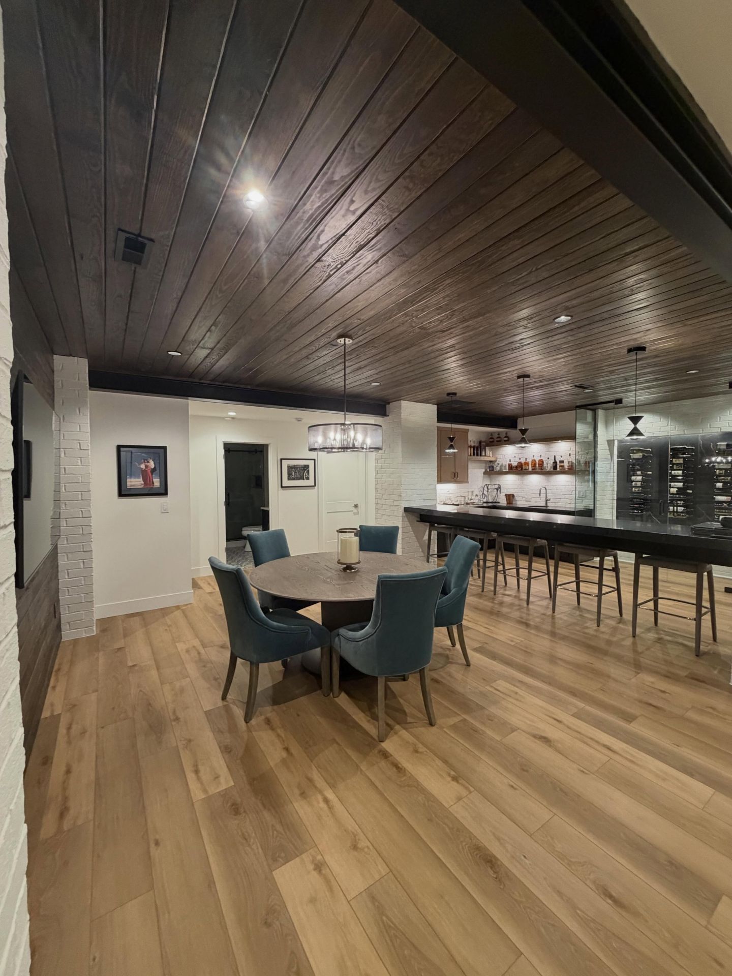 Empty modern lounge with wood floors, dark ceiling, round table, chairs, and bar stools by the counter.